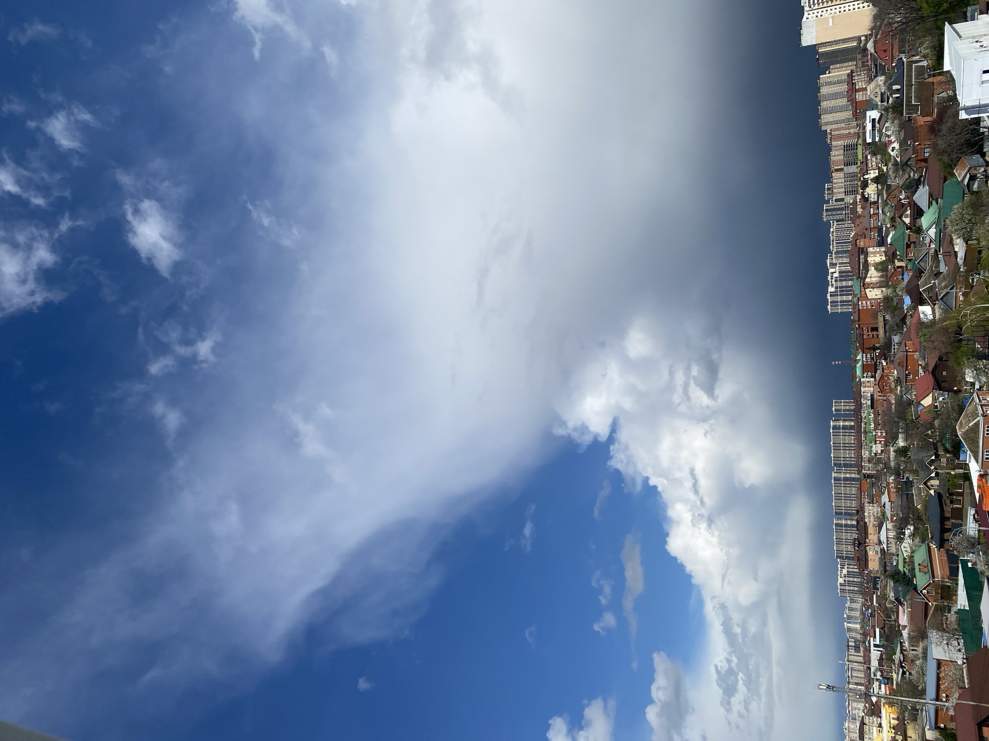 The image shows a view of an unidentified city. A clear, blue sky with scattered white clouds occupies the left side. On the right, dense, darker clouds appear, forming a sharp divide between the sky and the city buildings. The buildings consist of both residential houses with varying roof colors (red, green, brown) and taller apartment buildings. Trees are interspersed among the houses. The perspective is oriented such that the sky is to the left and the city is to the right. The location of the city is Unknown, Unknown.