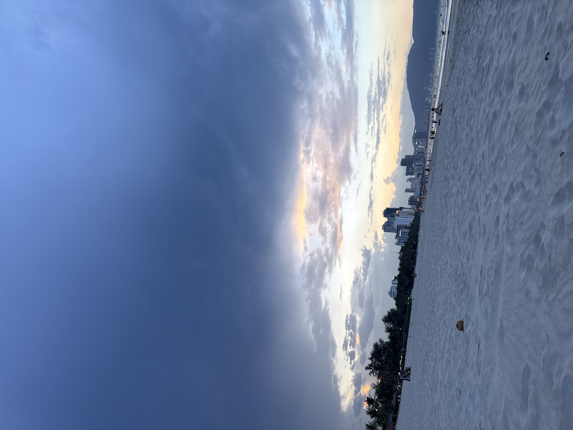 The image captures a panoramic view of a wide sandy beach at either sunrise or sunset, with a dramatic sky overhead. The left side of the sky transitions from a deep, clear blue to a vibrant display of clouds painted with warm hues of orange, pink, and yellow, interspersed with shades of grey and white, suggesting the golden hour.
A vast expanse of light-colored, fine-grained sand fills the right side of the frame, stretching from the foreground into the distance. The sand shows ripples and undulations. A single brown coconut rests on the sand in the mid-right. Along the horizon, a city skyline with numerous buildings and high-rises is visible, backed by distant, dark mountains. A line of green vegetation, including palm trees, fringes the edge where the beach meets the urban structures.
Several small figures of people are scattered along the beach, some near the water's edge, appearing as distant silhouettes. One more distinct figure is seen sitting or kneeling on the sand in the mid-ground. The overall atmosphere is serene and picturesque, highlighting the beauty of the transitioning daylight. No visible text is discernible in the image. The scene is set in Đà Nẵng, Vietnam.