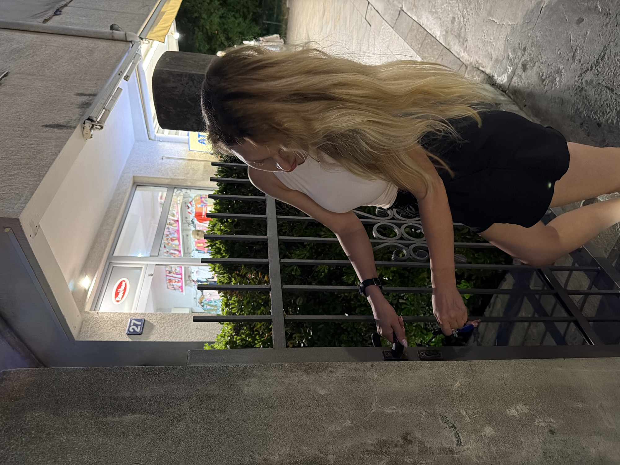 A woman with long blonde hair is using a key to unlock a metal gate in front of a building. She is wearing a white tank top and black shorts. The building appears to be a storefront with a sign that says "Rozeana" and the number 27. The scene is set at night, with artificial lighting illuminating the area. The background shows a street with a sidewalk and some greenery. The location is identified as Budva, Montenegro.