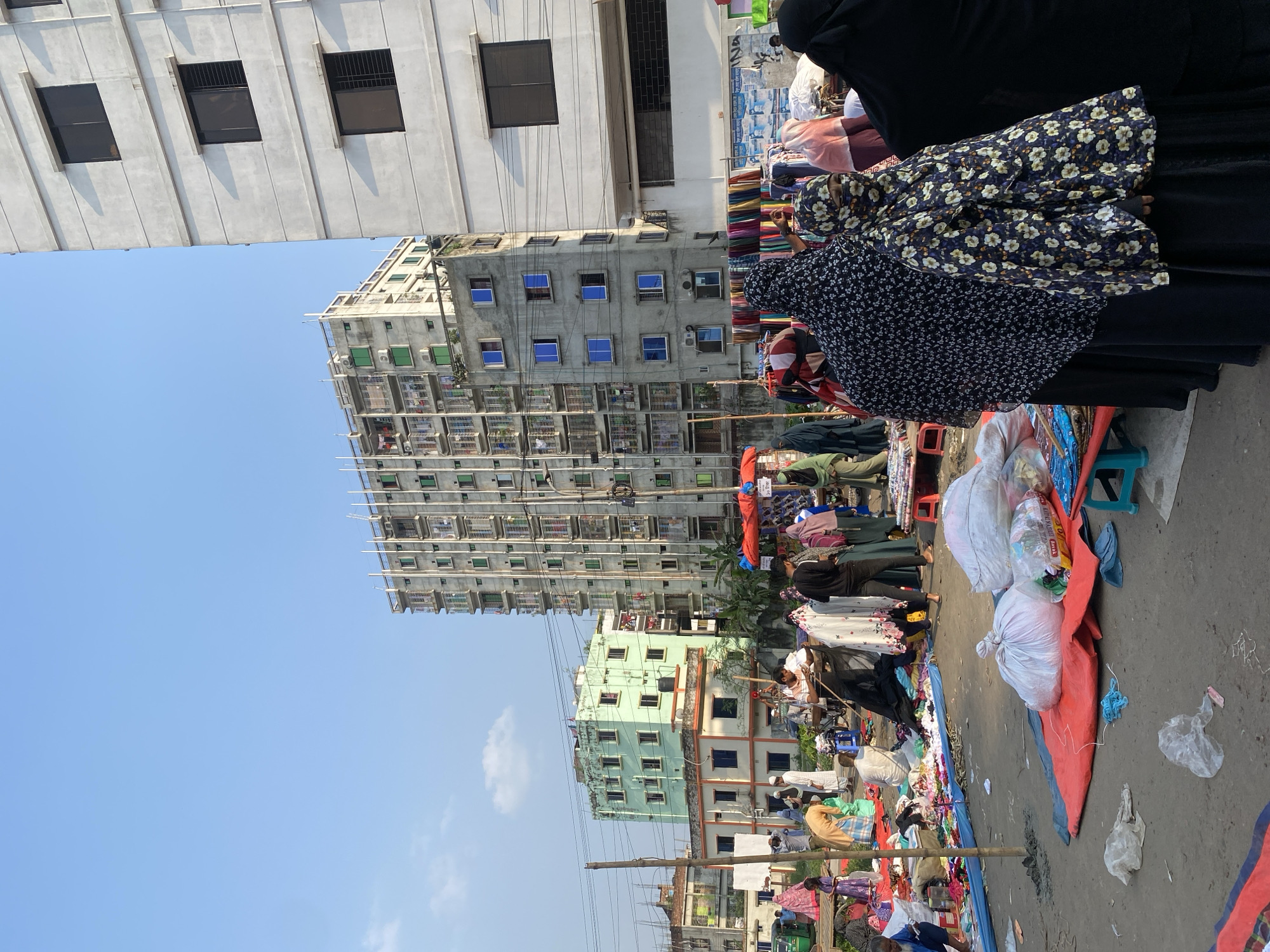 The image shows a bustling street market in Dhaka, Bangladesh. Several multi-story buildings dominate the background, one with a white facade and the others with varying shades of green and gray. In the foreground, vendors and customers are engaged in commerce, with clothing and textiles displayed on makeshift tables and tarps. Many of the women are wearing traditional Muslim attire, including hijabs and abayas. The street is crowded with people and goods, and the overall atmosphere is one of vibrant activity.