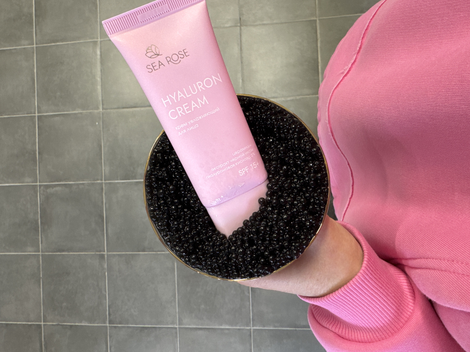 Here is a detailed description of the image:
The image shows a person holding a gold-rimmed dish filled with black beads. A pink tube of "Sea Rose Hyaluron Cream" is nestled within the beads in the dish. The tube is labeled "крем увлажняющий для лица" in Russian, translating to "moisturizing cream for the face". The label also states "экстракт черной икры гиалуроновая кислота 3% SPF 15+", meaning "black caviar extract hyaluronic acid 3% SPF 15+". The person is wearing a pink sweater. The scene is set on a grey tiled surface. There is no information about the city or location of the scene.