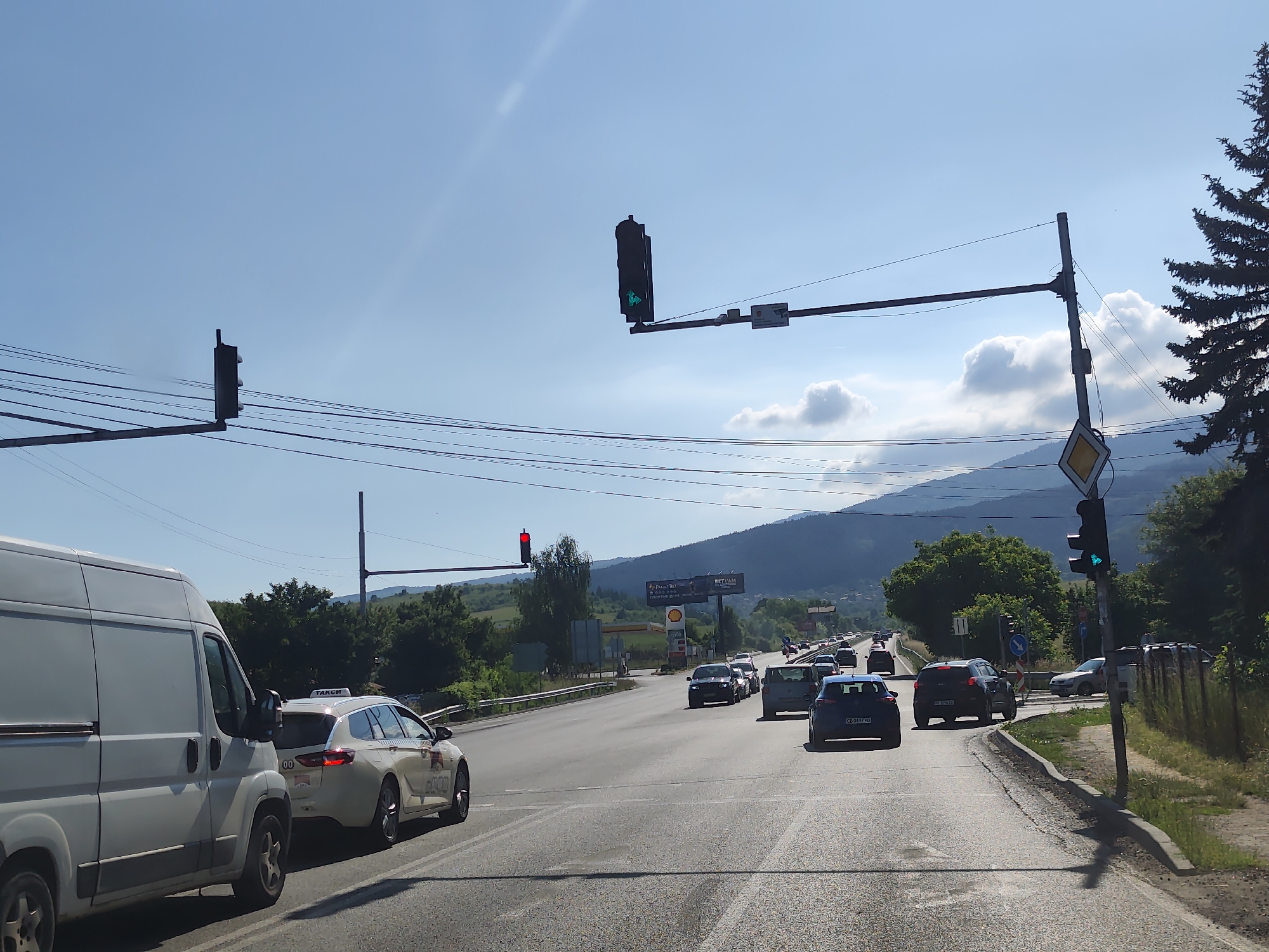 The image shows a street scene in Dragichevo, Bulgaria, under a clear blue sky with scattered clouds. Several cars are visible on a multi-lane road, some of which are stopped at a traffic light. A white van is in the foreground on the left, and a white taxi with "TAKSI" written on top is to its right. Further down the road, there is a traffic light showing red, and another traffic light on the right side of the road is showing green with a left-turn arrow. In the background, there are green hills and mountains, trees, and a billboard advertising "BILLA" and "SHELL". The scene appears to be a typical road intersection on a sunny day.