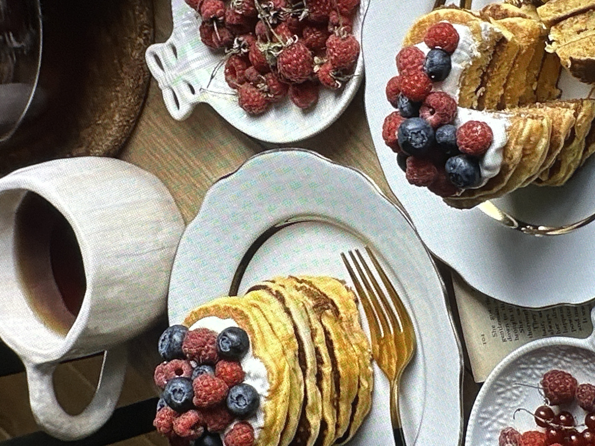 A high-angle, still-life image depicts a breakfast or brunch scene. A white, textured ceramic mug filled with a dark liquid, likely tea or coffee, sits on the left. Adjacent to the mug, a white plate with a gold rim displays a stack of pancakes layered with cream and topped with blueberries and raspberries. A gold-colored fork rests on the plate next to the pancakes. In the top center of the image, another white plate holds a pile of raspberries. A third plate to the right features a similar pancake stack with berries and cream. The entire scene is set on a textured surface; fragments of a book are visible near the bottom right. No people are directly visible in the image. The setting appears to be indoors, perhaps a cafe or restaurant. The city of Yerevan, Armenia is not discernible.