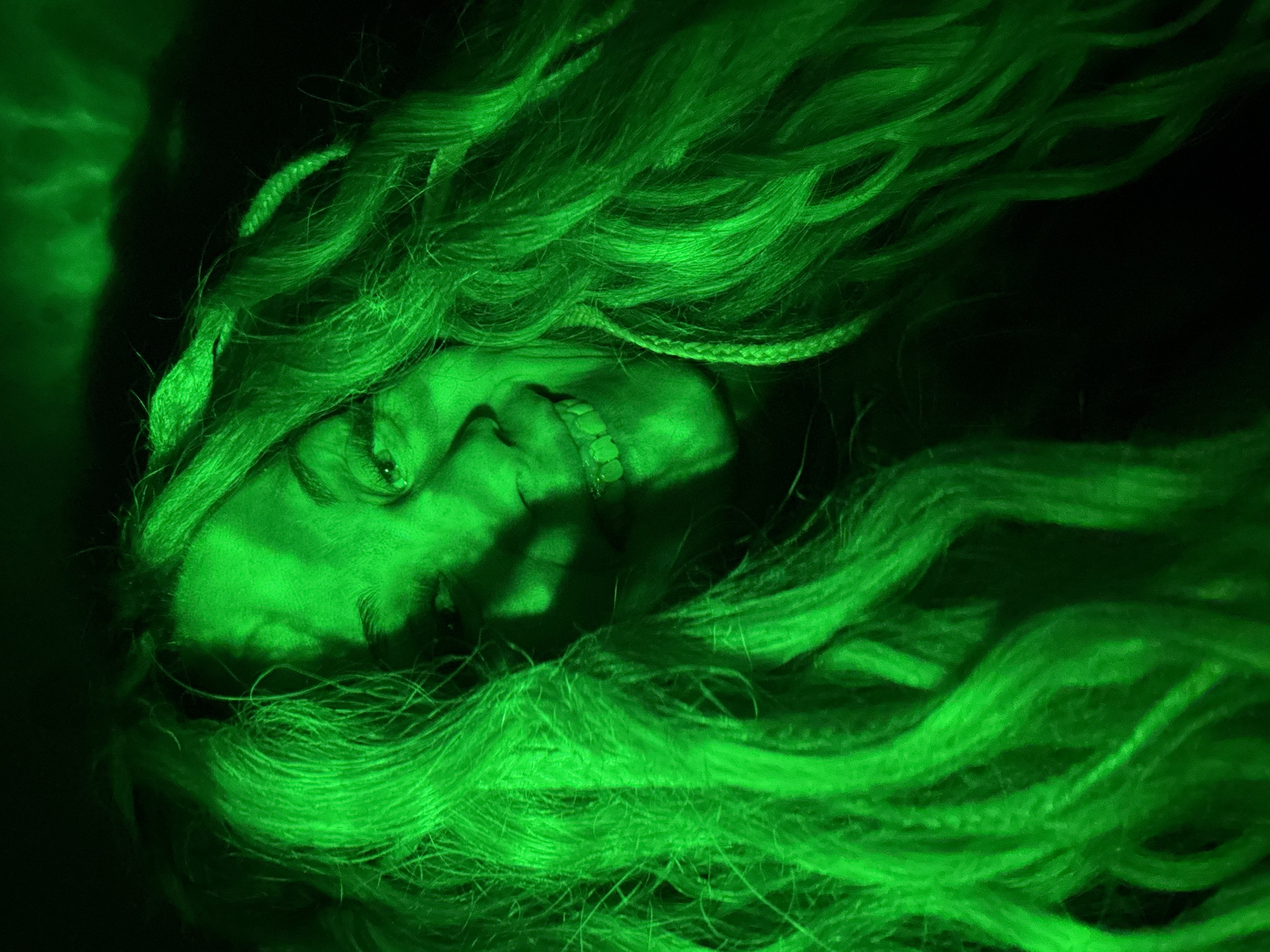 A person with light skin and long, light-colored hair is illuminated by green light. The person is lying down with their face tilted upwards and a slight smile visible. Braids are present in the hair. The background is dark and indistinct. There is no evidence to suggest the image was taken in Rusanovo, Russia.
