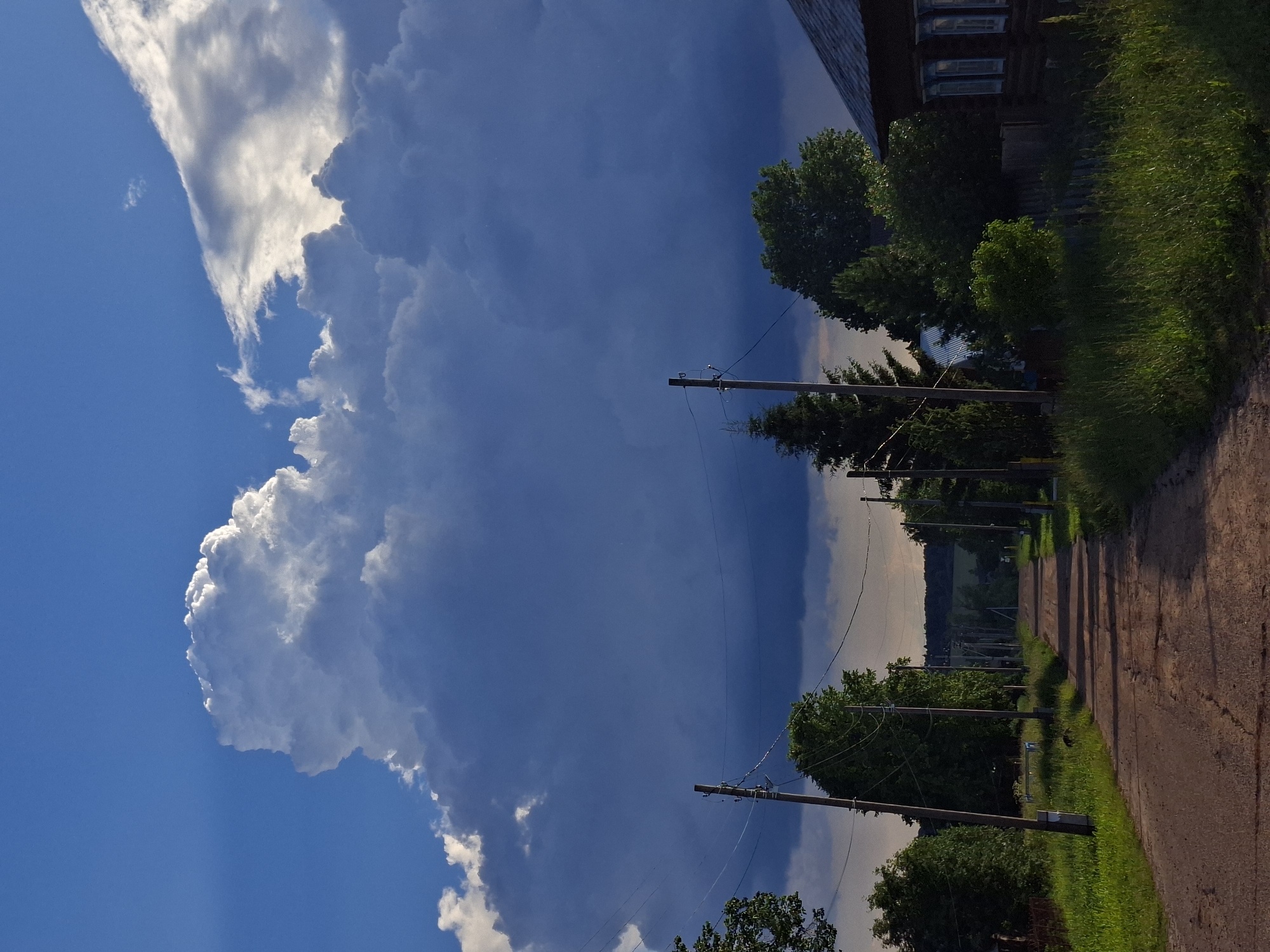 The image depicts a rural road in a village, possibly in Eastern Europe, under a dramatic sky. A dirt road stretches into the distance, flanked by green grass and trees. Utility poles with wires line the sides of the road. On the right, a wooden house with blue window frames is visible. The sky is dominated by a large, dark storm cloud, with a bright, illuminated cloud formation on its left. The scene suggests an approaching storm or the aftermath of one. There are no people or animals visible in the image. The time of day appears to be late afternoon or early evening, given the quality of light.