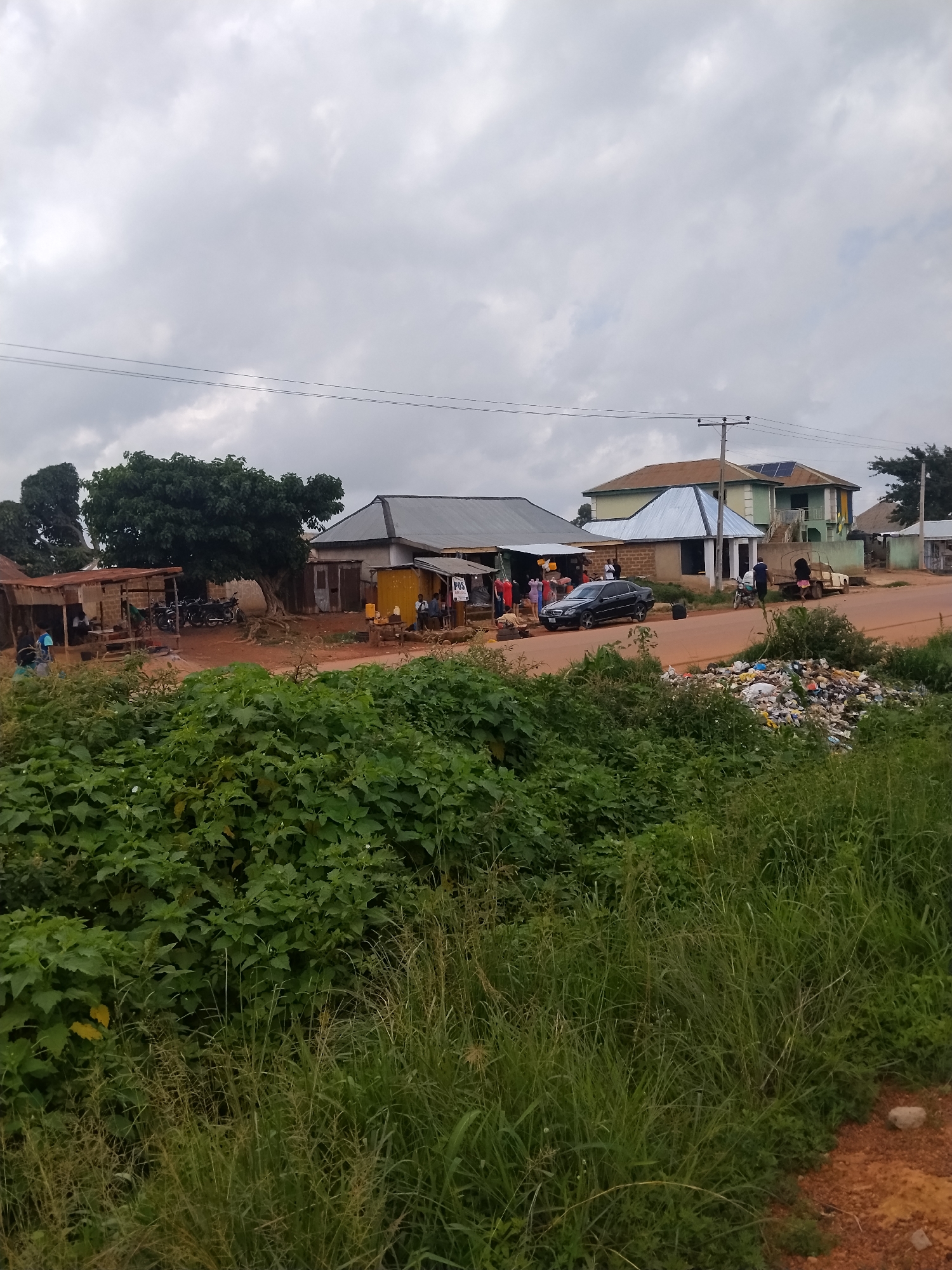 The image depicts a street scene in Durumi, Nigeria, with a focus on a roadside market and residential buildings under an overcast sky. In the foreground, lush green vegetation and tall grass line the edge of a dirt road. The middle ground reveals a collection of market stalls with corrugated metal roofs, some constructed with brick and wood. Several motorcycles are parked near one of the stalls. A black sedan is parked on the roadside in front of another building with a light blue metal roof. People are visible interacting at the market stalls, some appearing to be customers and vendors. Further back, residential buildings with painted exteriors and metal roofs are present, with one prominently featuring solar panels on its roof. To the right of the road, a large pile of refuse is visible, contributing to a sense of informal waste management. The overall impression is of a busy, everyday street life in a Nigerian community, characterized by a mix of commercial activity, residential structures, and natural elements.