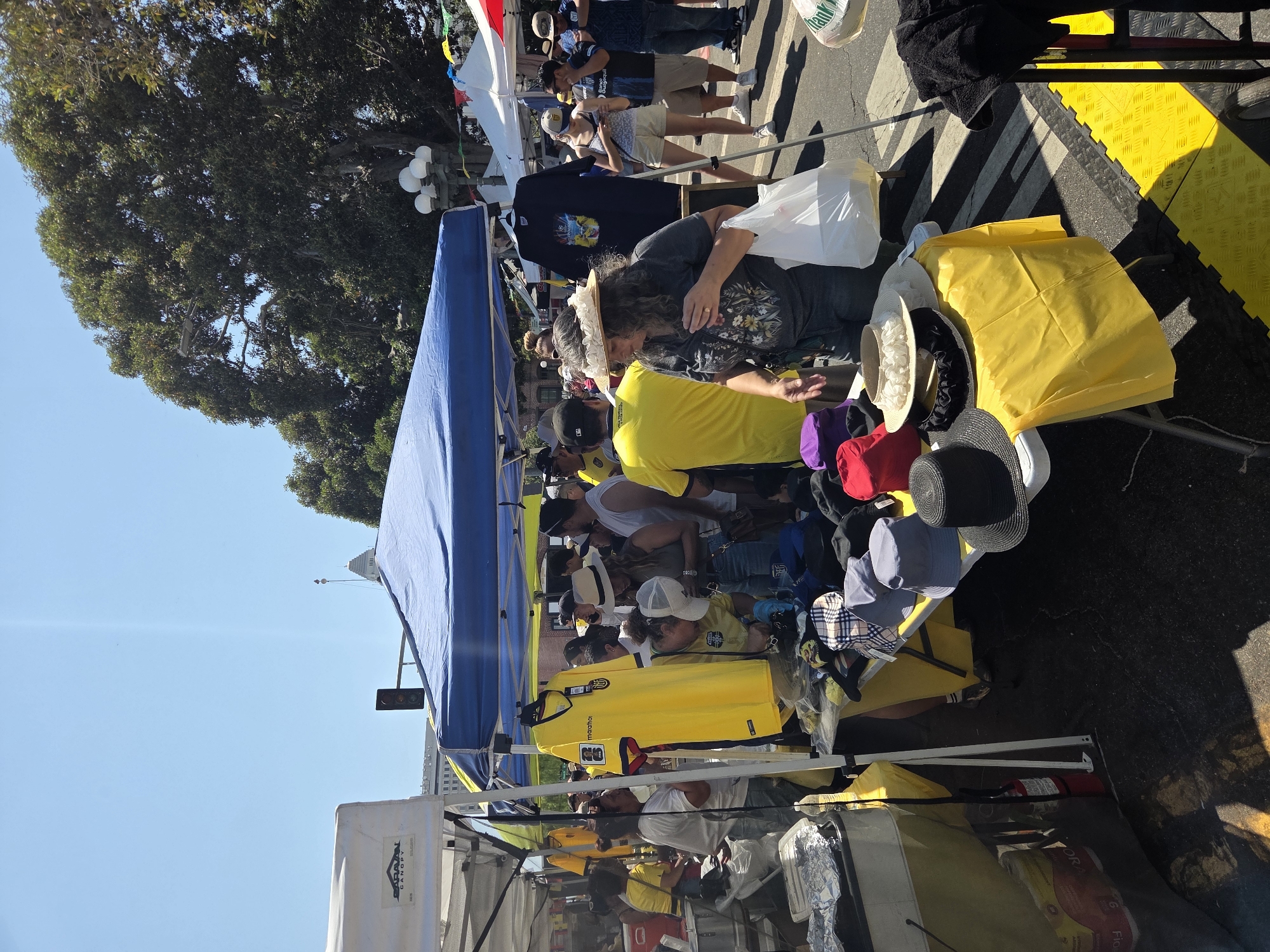 This image depicts an outdoor market scene, likely in Los Angeles, USA, given the presence of large trees and a clear, sunny sky. The foreground is dominated by a table laden with hats of various styles and colors, including fedoras, bucket hats, and a wide-brimmed straw hat adorned with ruffles. A woman with curly hair, wearing a dark t-shirt and a decorative straw hat, is seen holding a white plastic bag and looking down at the table. Behind her, a vibrant yellow t-shirt is prominently displayed on a hanger, possibly indicating merchandise for sale.
Several people are browsing or standing around the market stalls, many wearing casual attire. Among them, individuals are dressed in yellow shirts, suggesting a potential theme or affiliation. Several market tents, one blue and one white, are visible, providing shade and housing the merchandise. In the background, a traffic signal is visible, and glimpses of buildings and lush green trees indicate an urban environment. The ground appears to be asphalt with yellow and black patterned mats laid out in some areas, possibly for pedestrian traffic or aesthetic purposes. The overall atmosphere suggests a bustling event or street fair.
