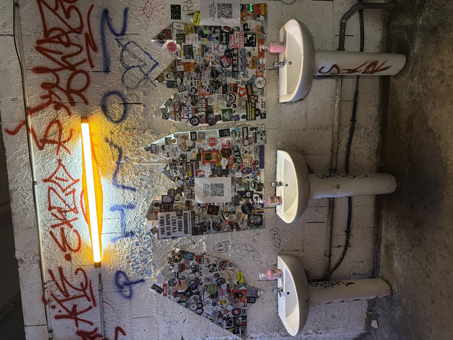 This image depicts an interior wall section of a public facility in Bagnolet, France, likely a washroom. The textured, white-painted wall is heavily covered in red and blue spray-painted graffiti tags and stylized lettering. A single, horizontally mounted fluorescent light fixture, emitting an orange glow, is positioned centrally on the upper wall. Below the light, three irregularly shaped panels or sections of the wall are densely overlaid with a multitude of diverse stickers, some containing text such as "PAS SÛR-E DE CE QU'ON T'A FILÉ? PENSE À MICRODOSER" and QR codes. Three white ceramic pedestal sinks are arranged in a line along the lower portion of the wall. Each sink features a single metal faucet and a pink or translucent pink plastic pump bottle, presumably containing soap or sanitizer, resting on its basin. The pedestals of the sinks display minor graffiti. Exposed dark-colored pipes run horizontally along the wall behind the sink pedestals, near the concrete floor, which shows signs of wear and staining. No individuals are present in the scene.