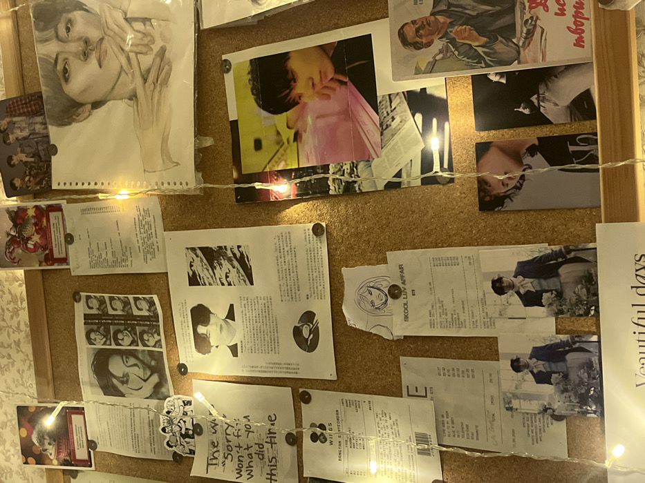 The image displays a cluttered cork board, situated on a wall, depicting a personal collection of memorabilia within a residence in Chaikovsky, Russia. The board is densely covered with photographs, printed papers, drawings, and notes, secured by small, dark-colored pins, some appearing to be coins. A strand of warm white LED string lights is draped vertically across the central portion of the board, providing illumination.
On the upper left, a greeting card features a furry animal in a party hat, partially obscured Russian text visible below. Beneath this, a white paper shows a large image of a woman's face next to a grid of smaller portraits of a man. A black-and-white sticker depicting cartoon figures is also present.
The central-left area includes a handwritten note on white paper, stating, "The worst sorry won't fix what you did this time." Below this are two printed documents resembling music album tracklists or receipts; one lists songs from "BTS WINGS," and the other is titled "SKOOL LUV AFFAIR BTS." Two small, identical rectangular photographs below these display a man, identified as a K-pop idol, in historical-style attire, holding flowers. A partially visible white paper at the bottom reads "Veautiful days."
Towards the upper center-right, a small photograph depicts a K-pop group. Above it, a red Christmas-themed greeting card is visible. A prominent white paper in the central section features a black-and-white portrait of a man, identified as a K-pop idol, at the top, flanked by columns of Japanese text and an abstract graphic, with a vinyl record graphic at its base.
The right side of the board exhibits a large, realistic pencil drawing of a man, identified as a K-pop idol, with one hand near his chin, partially covered by clear plastic. Below this, a vibrant color photograph features another man, identified as a K-pop idol, with dark hair, leaning contemplatively on a purple surface against a yellow background. A small white paper with a minimalist line drawing of a woman's face is pinned nearby. Further right, a poster in a Soviet-era style portrays a man in work clothes smoking, with Cyrillic text visible underneath. Two vertical photographs of men, identified as K-pop idols, one partially revealing "Calvin Klein" branding and the other showing a lower torso in jeans, are located at the bottom right.
The wall directly behind the cork board appears to be covered with light-colored, textured wallpaper.