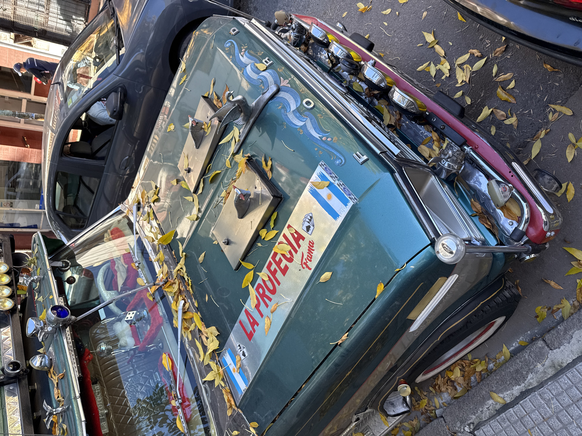 The primary subject is a highly customized, vintage-style vehicle, possibly a car or small "colectivo" (bus), painted teal green with a red roof trim and decorative accents. It's adorned with various chrome fixtures, including a bird-shaped hood ornament and numerous auxiliary lights, some with yellow lenses. A dark grey modern car is parked alongside it, with a small portion of another dark vehicle visible in the upper right. One person in a dark uniform (blue cap and jacket) is seen standing near the grey car in the background. Yellow autumn leaves are scattered across the customized vehicle and the asphalt street.
The vehicles are parked on a city street. No specific activity or event is taking place beyond this static scene.
Notable details include the elaborate decoration of the teal vehicle. Its hood features intricate blue and white wavy paint designs, reminiscent of the Argentine flag colors, and multiple decals of the Argentine flag (including the sun emblem) are visible. The car has classic whitewall tires. Through the windshield, a small disco ball and a red, possibly fluffy, steering wheel cover are visible inside. The abundance of yellow leaves suggests an autumn season. The scene is brightly lit, indicating daytime. The unique customization style of the vehicle, particularly if it's a "colectivo," is a cultural hallmark of Buenos Aires, Argentina.
Visible text includes: "LA PROFECÍA" in prominent red letters, with "Fama" printed below it on a decal. Small "Ford" logos are identifiable on some chrome parts, such as a hubcap or a decorative element near the front wheel. A partial inscription "Club Amigos del..." can be seen on the side of the teal vehicle. Another sticker displays an image of playing cards, specifically two aces and two kings.