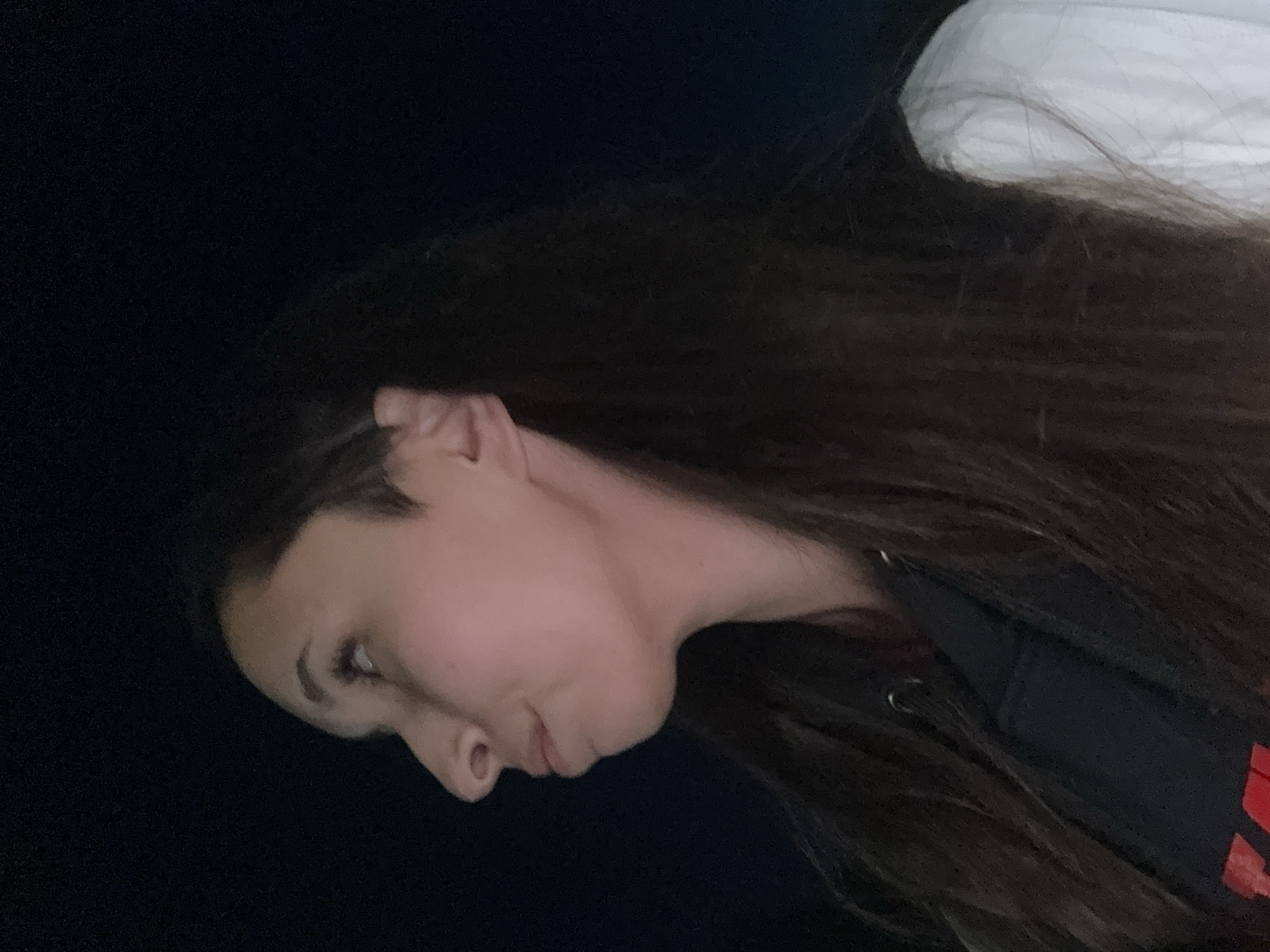 A close-up, profile view captures a young woman with long, dark hair looking upwards and to the left. She is wearing a black hooded sweatshirt and a white garment over it. The background is dark and featureless, suggesting an outdoor setting at night. The image appears to be taken in Doma 1096 Km, Russia.