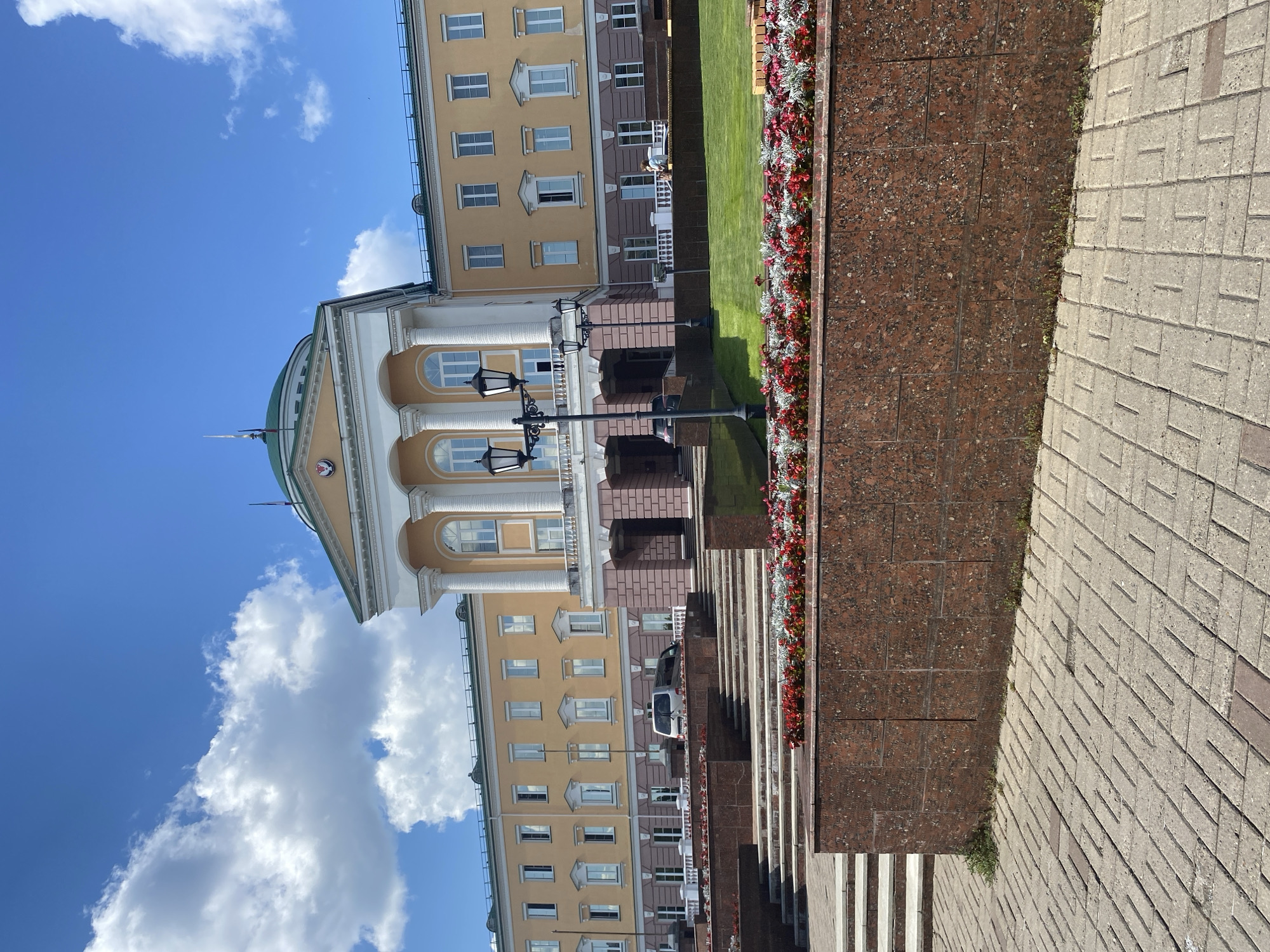 This is a daytime, exterior photograph taken in Izhevsk, Russia. The image features a large, multi-story building with a light yellow facade, white columns, and arched windows. The building has a green dome and a flagpole. In front of the building, there is a flowerbed with red and white flowers, followed by a granite-tiled walkway and then a brick-paved area. The sky is bright blue with fluffy white clouds. The scene appears to be a public space, possibly an administrative or historical building. There are no people clearly visible in the frame, but a white van is parked near the building in the distance. The overall impression is one of a sunny day in a well-maintained urban environment.