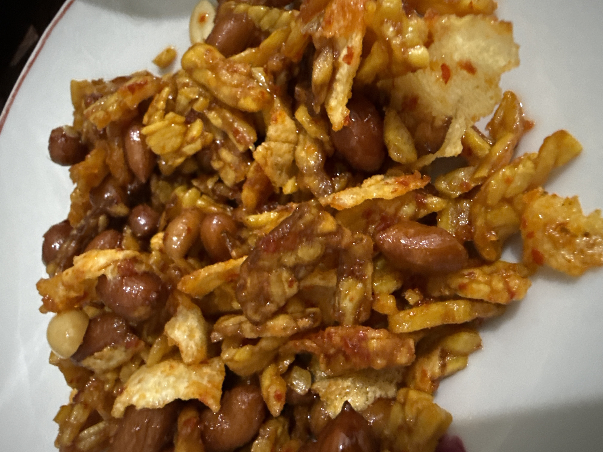 The image shows a close-up, overhead shot of a plate of food. The food appears to be a mix of fried tempeh, peanuts, and possibly crispy potato slices, all coated in a glossy, reddish-orange sauce with visible chili flakes. The dish is piled high on a white plate with a thin red rim. The lighting suggests indoor illumination. The context provided regarding Lhokseumawe, Indonesia, is external to the visual information and cannot be corroborated from the image itself.
