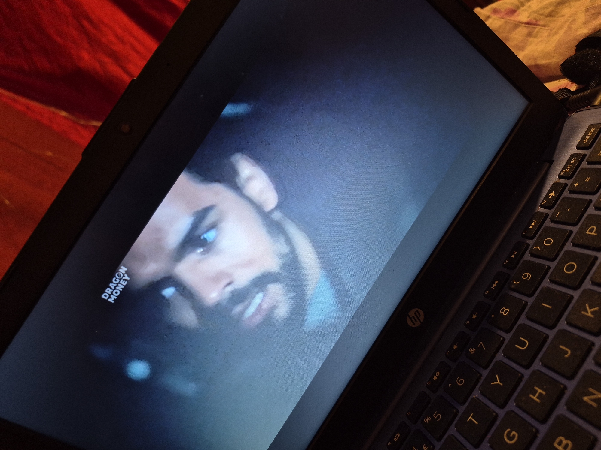 The image shows a close-up view of a laptop screen displaying a film or video. The screen shows a man's face, with visible blue eyes and a beard. The words "DRAGON MONEY" are displayed in white text at the top of the screen. The laptop is open, and a portion of the keyboard is visible at the bottom of the image. The laptop is placed on a surface that appears to be a red fabric. There are no identifiable landmarks or details that would indicate a specific location within Sandwell, United Kingdom. The scene is indoors, likely in a domestic setting, with no external environmental factors visible.