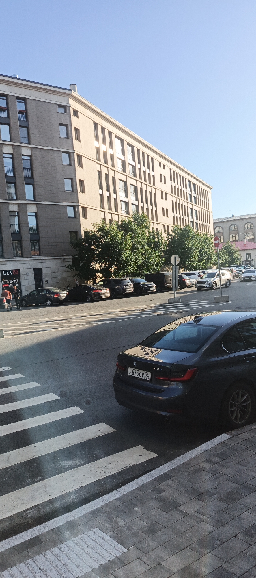 A street view in Saint Petersburg, Russia, shows a multi-story building with numerous windows and balconies. A pedestrian crossing with white stripes is visible in the foreground. Several cars are parked along the street, including a dark sedan with a license plate that reads "H675OY 75" and a white SUV. Trees line the sidewalk in front of the building. The sky is clear and blue.