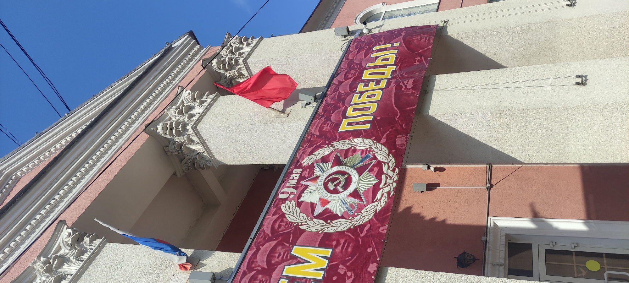 The image depicts a building in Fryazino, Russia, adorned with decorations commemorating Victory Day on May 9th. A large banner hangs vertically on the building’s façade, displaying a Soviet-era emblem of the Order of the Patriotic War, along with the text “9 мая” and “Победа!” (Victory!). Red and Russian flags are mounted on the building’s exterior, adding to the festive atmosphere. The building itself features architectural details such as decorative molding and arched windows, suggesting a historical or administrative function. The sky is clear and blue, indicating fair weather conditions.