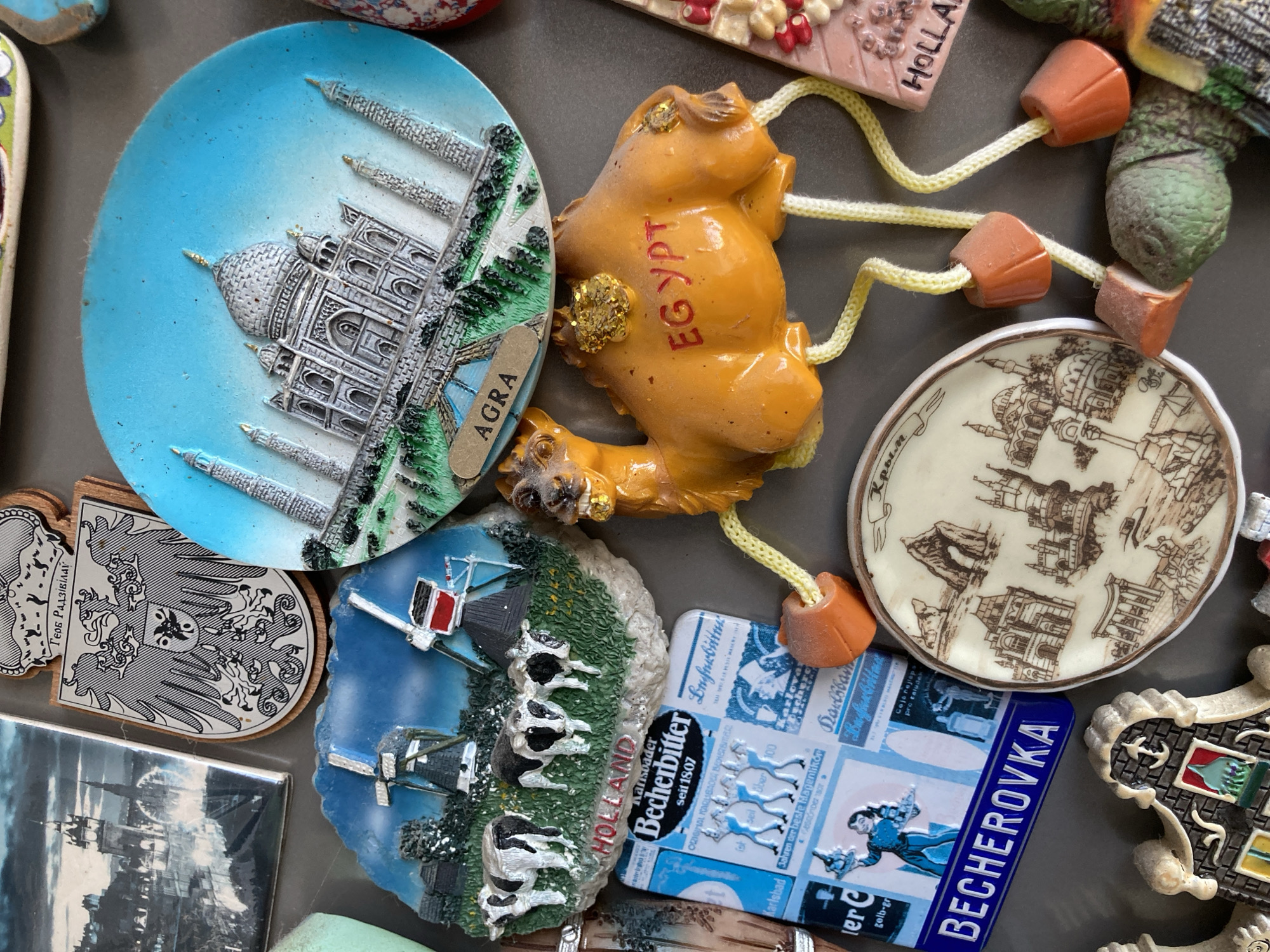 A collection of novelty refrigerator magnets is arranged on a dark gray surface. In the upper left is a magnet featuring a black and white image of a historic European city with a bridge and tower. To its right is a magnet with a shield bearing a double-headed eagle and a crown, with text in Cyrillic. Below these are two round magnets: one depicts a blue sky with a detailed 3D representation of the Taj Mahal in Agra, India, labeled "AGRA"; the other features a 3D scene of Dutch windmills and cows with the word "HOLLAND" visible.
Dominating the center of the image is a bright orange, three-dimensional camel magnet, with yellow glitter on its humps and red lettering on its body that reads "EGYPT". The camel has four cord legs with orange stoppers. To the left of the camel magnet is a rectangular magnet depicting a scene of windmills and cows, with the word "HOLLAND" in red letters below. Below this is a blue and white rectangular magnet for "Becherovka" liqueur, featuring historical images and text.
To the right of the camel, partially visible, is a pink rectangular magnet with colorful mosaic-like decorations and the word "HOLLAND" in black letters. Below this and to the right is a round magnet with a sepia-toned drawing of various architectural structures, possibly castles and domes, and text in Cyrillic. In the bottom left corner is a magnet shaped like a building facade with intricate details. A green, textured magnet resembling a creature or plant is visible in the bottom right. The overall arrangement suggests a display of souvenirs from various international locations.
