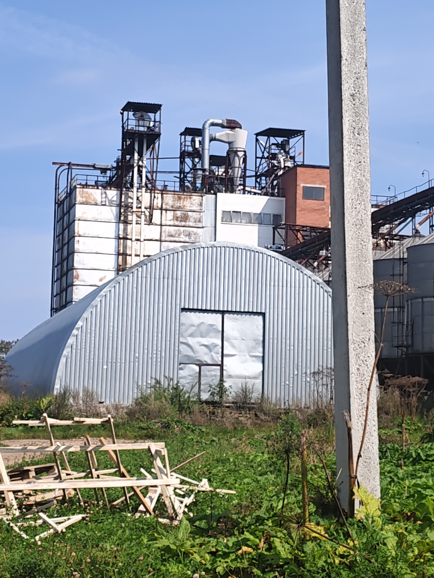 A low-angle, vertical shot captures a section of an industrial facility on a clear day. In the foreground, a concrete pillar stands to the right, partially obscuring the view of the plant. In front of the pillar and the industrial structures, a collection of weathered wooden elements, possibly scrap materials or construction remnants, is scattered amidst lush green vegetation.
Behind the foreground elements, a large, silver, arched building dominates the mid-ground. This structure features a corrugated metal exterior and a large, metal-clad doorway with two panels. To the left of this arched building, a taller, white industrial silo or structure rises, showing signs of weathering and rust. This silo is part of a larger complex that includes metal platforms, scaffolding, pipes, and a red brick building with a window. The top of the silo complex features open-air platforms with metal roofs and railings.
The background is a clear blue sky. The overall impression is one of an operational or disused industrial site set against a natural, overgrown landscape. The location is identified as Platoshino, Russia. There are no people visible in the image.