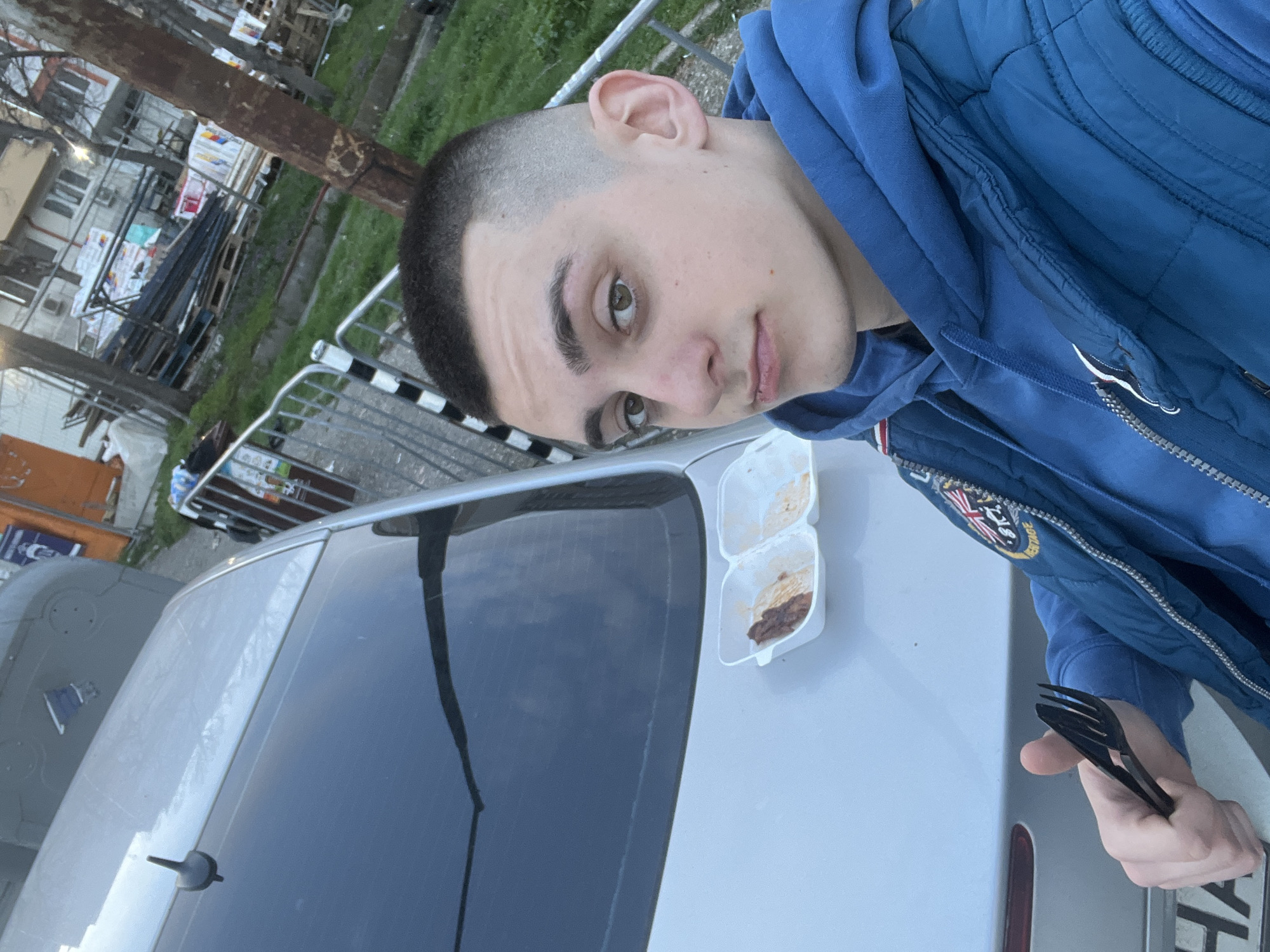 The photograph shows a young man with short, dark hair standing next to a silver car, possibly in an urban area. He is wearing a blue hooded sweatshirt and a blue jacket with a patch on the sleeve, holding a black plastic fork in his right hand. On the car's trunk, there's a partially-eaten meal in a white styrofoam container. The background includes buildings, a tree, and a metal fence, suggesting an outdoor setting in Burgas, Bulgaria.
