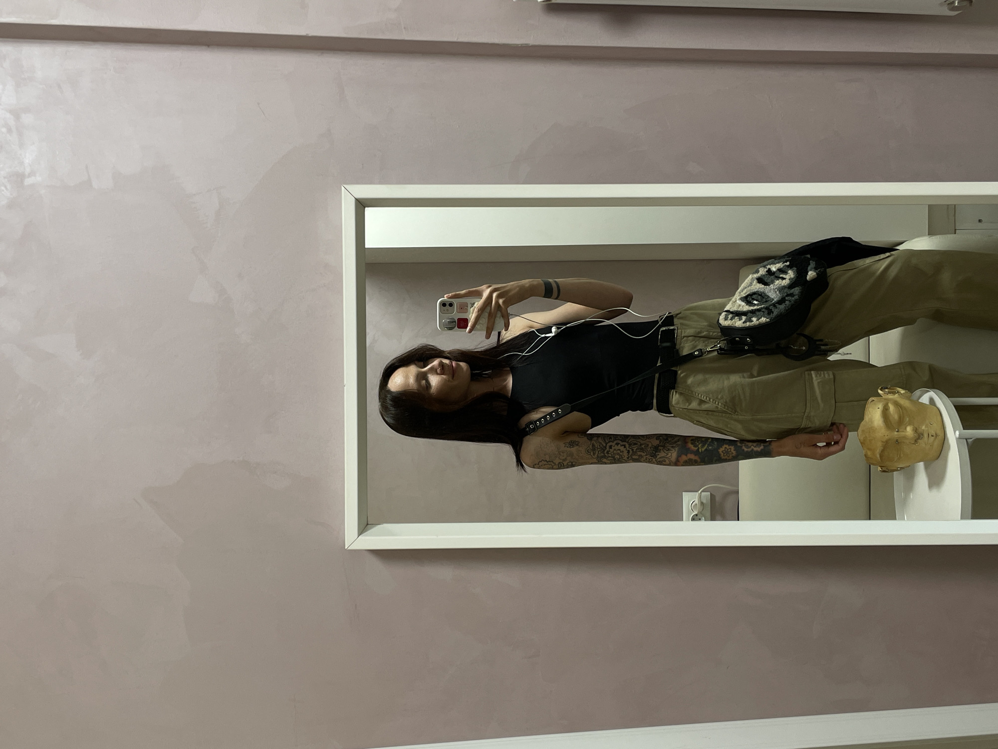 A woman is taking a selfie in a mirror, presumably in Izhevsk, Russia. The mirror is framed in white and reflects the woman's upper body and part of the bathroom. She is wearing a black top, khaki pants, and a skull patterned bag. She has tattoos on her arm. A gold-colored mask is on the edge of a bathroom sink. The wall behind the mirror is a mottled pinkish-beige color.