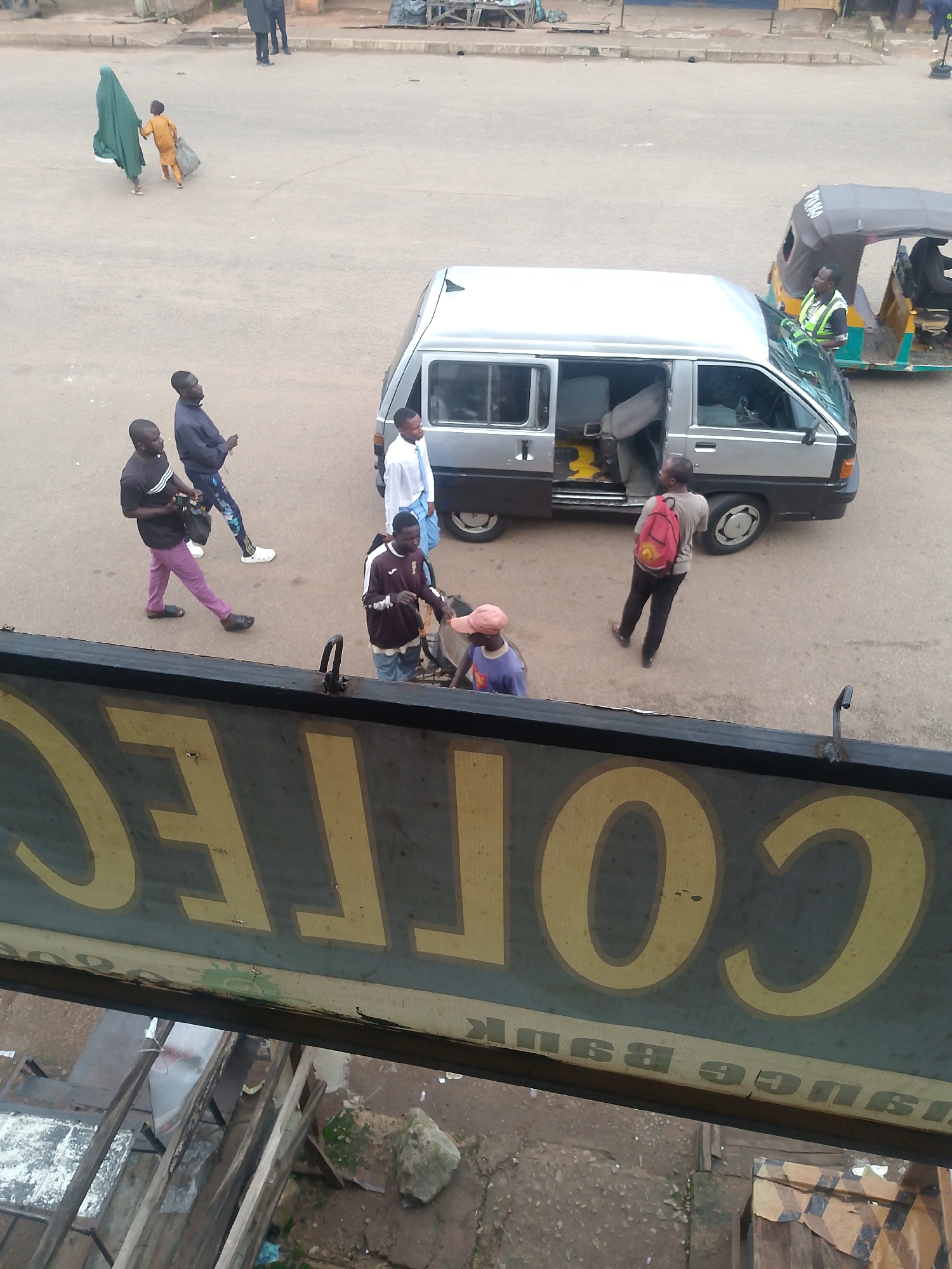 The image captures a street scene in Bukuru, Nigeria. In the foreground, a section of a sign with large yellow letters, possibly reading "COLLEGE" and "INSURANCE BANK" (partially obscured and reversed), is visible. Beyond the sign, a silver minibus with open doors is parked on the paved road. Several individuals are near the minibus. One person in a dark shirt and purple pants is walking away from the viewer. Another person in a dark jacket and patterned pants is walking past the minibus. A third person in a maroon sweatshirt and blue jeans is standing next to a bicycle. A young person wearing a blue shirt and a pink hat is also present. A person wearing a red backpack is standing near the minibus, facing away from the viewer. In the middle ground, a yellow and grey auto-rickshaw is driving away from the camera. Further in the background, two people, one in a green headscarf and the other in a yellow garment, are walking on the street, with the person in green holding a dark bag. The environment appears to be an urban setting with a paved road and buildings in the distance.