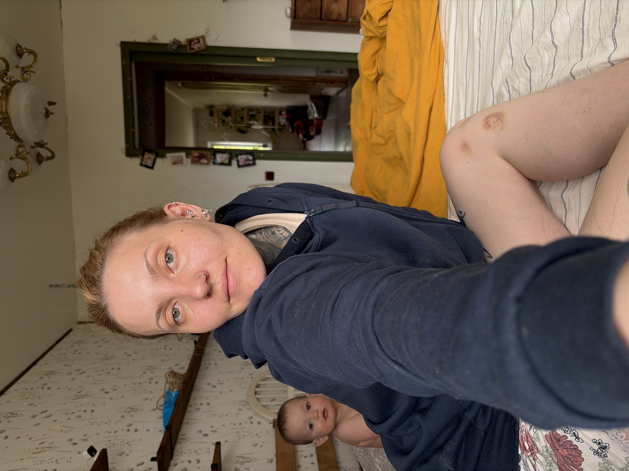 A selfie-style photograph shows a light-skinned woman with blonde hair pulled back and numerous piercings in her left ear. She is wearing a dark blue hooded sweatshirt and appears to be lying on a bed. A baby is partially visible beneath her, lying on the same bed. The woman has bruises on her leg. The bed has striped linens and a yellow blanket. The room is cluttered, with a mirror on the wall adorned with small photographs, a light fixture on the ceiling, and patterned wallpaper. The location is Chaikovsky, Russia.