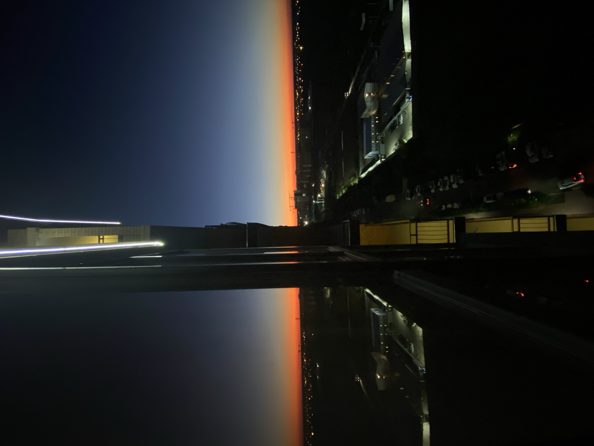 The image captures a twilight view from a high-rise building in Krasnodar, Russia. The sky transitions from a deep blue at the top to a vibrant orange and red near the horizon, indicating sunset or sunrise. In the foreground, the reflective surface of the building's exterior creates a mirror image, splitting the scene vertically. On the right side of the reflection, balconies with yellow railings are visible.
Beyond the building, the cityscape is illuminated by streetlights and lights from buildings, suggesting it's dusk or dawn in an urban or semi-urban area. Several industrial-looking buildings with flat roofs are present, some with parking lots. Cranes are visible in the distance, hinting at ongoing construction. A street with parked cars runs along the lower part of the image. The overall atmosphere is calm and serene, with no people or specific activities clearly identifiable. The focus is on the urban landscape against the backdrop of a colorful sky.