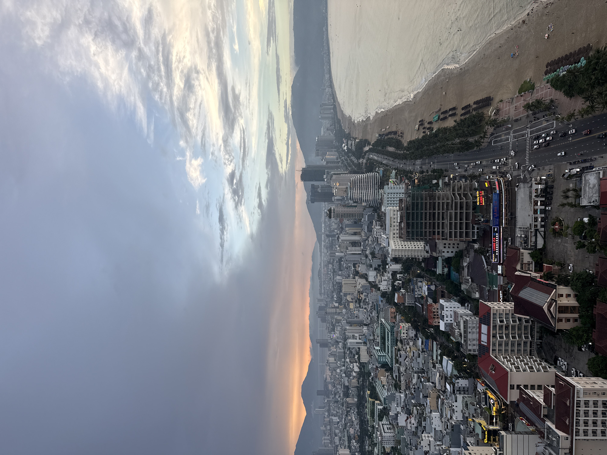 This image captures an aerial view of the coastal city of Đà Nẵng, Vietnam, likely taken at sunset. The scene is divided into distinct zones: the city skyline, a beach, and the sea.
In the foreground and mid-ground, a dense urban landscape stretches inland. Numerous buildings of varying heights and architectural styles dominate this area, with a mix of residential and commercial structures. Some buildings are modern high-rises, while others appear older and more traditional with red-tiled roofs. Roads are visible, with cars and motorbikes navigating through the city. Streetlights and building lights are beginning to illuminate, suggesting dusk.
Adjacent to the city is a long, sandy beach. Palm trees line the edge of the beach, and rows of umbrellas and lounge chairs are set up, indicating a popular tourist destination. A number of people are scattered on the beach, some walking along the shore, others relaxing under the umbrellas. The sea meets the beach with gentle waves.
In the background, a range of mountains form a natural backdrop to the city, their peaks shrouded in a soft haze. The sky above is a dynamic mix of dramatic clouds. On the left side, the sky is filled with heavy, dark grey clouds, hinting at recent or approaching rain, with a soft orange glow visible on the horizon between the city and the mountains, indicative of sunset. On the right side, the sky transitions to lighter blues and whites, with wispy, textured clouds catching the fading sunlight, creating a more serene atmosphere. The overall lighting suggests the transition from day to night, with a warm, soft glow illuminating the scene.