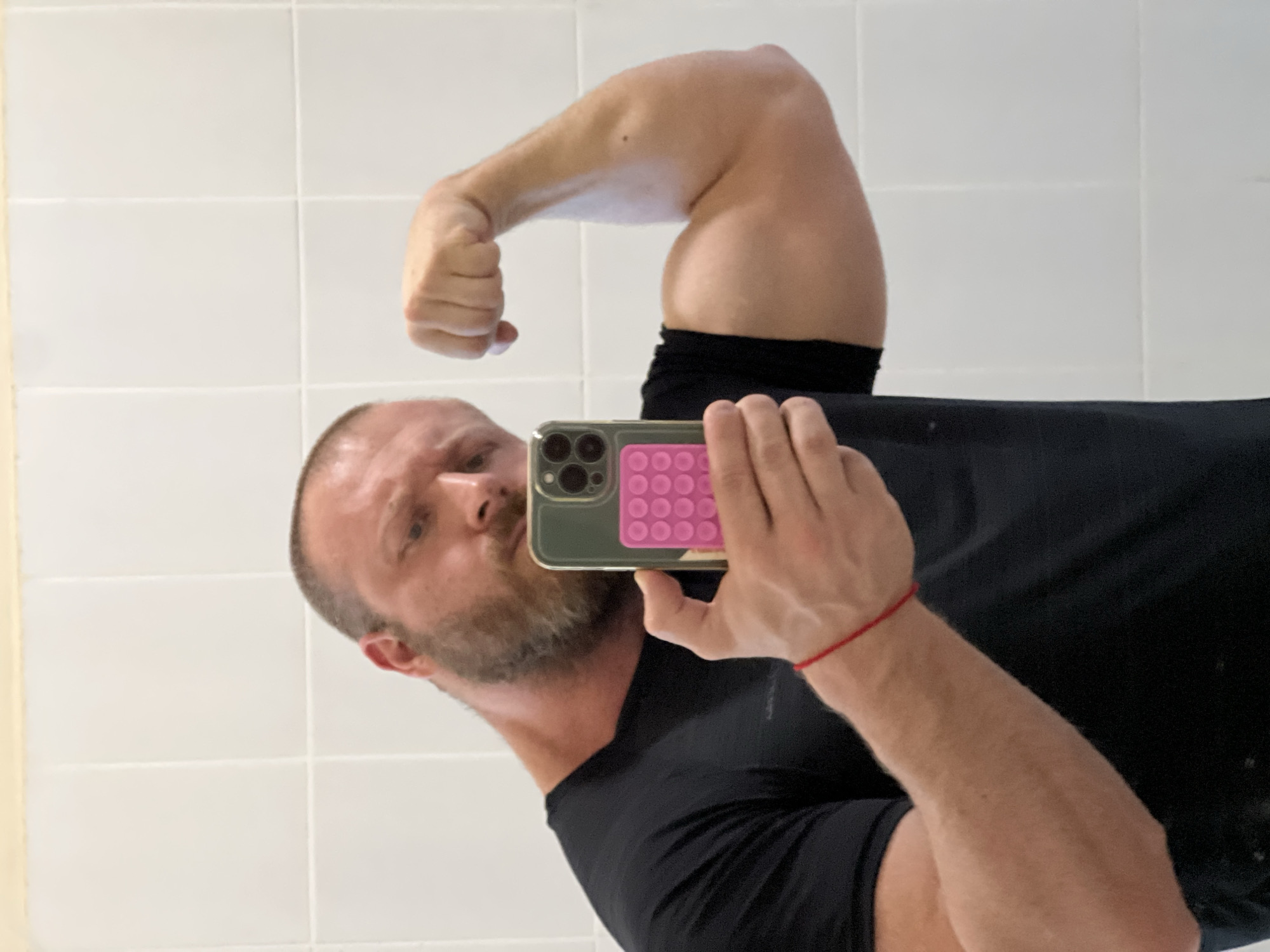 A man with a beard and a shaved head is taking a selfie in a bathroom. He is wearing a black t-shirt and flexing his left bicep. He is holding a smartphone with a green case with a pink pop-it on the back. The bathroom has white tiled walls. There is no indication of the location being Chaikovsky, Russia.