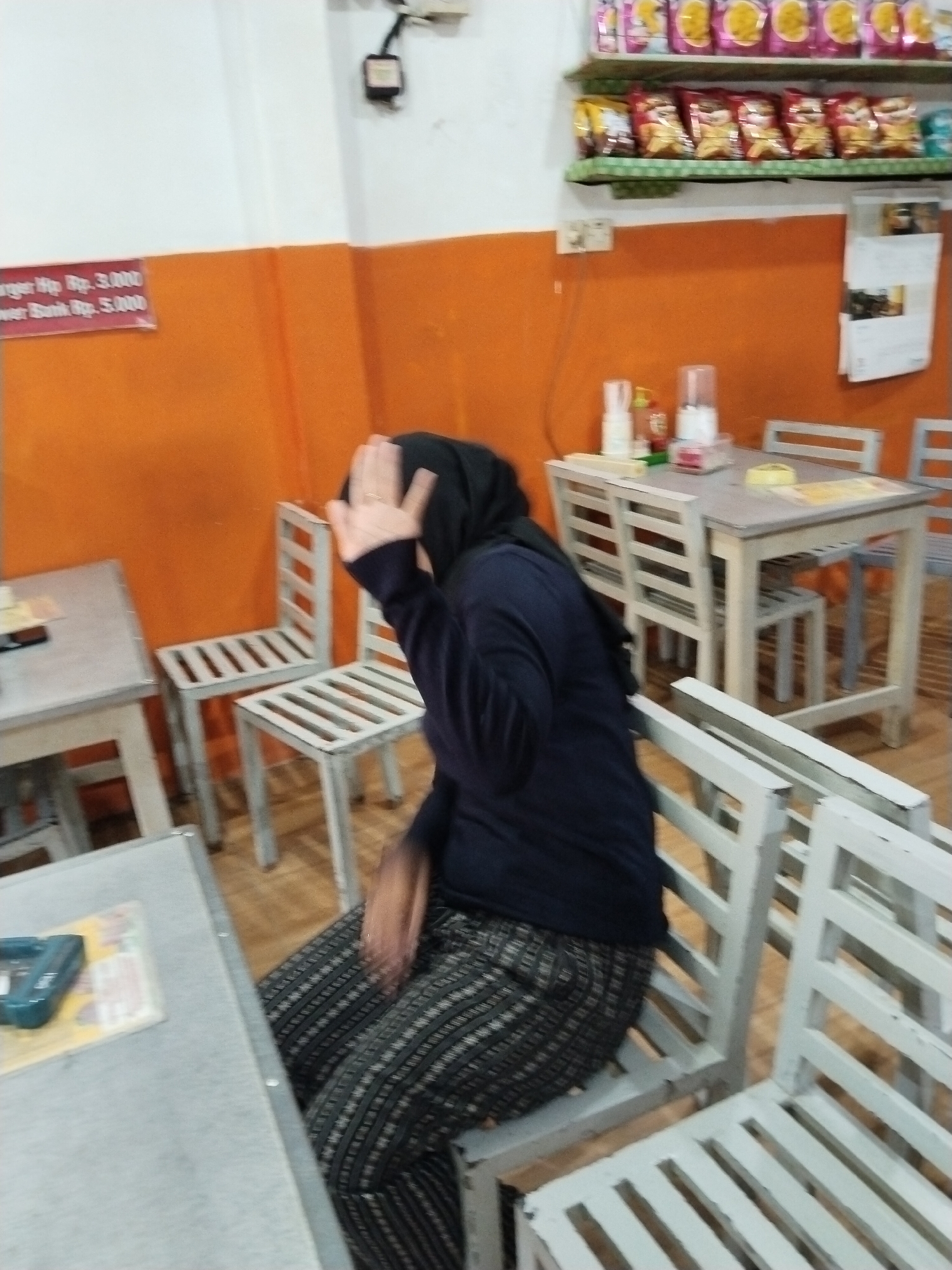 A person wearing a black headscarf and a dark blue top is seated at a table in what appears to be a restaurant or cafe in Prigi, Indonesia. The person is partially obscuring their face with their right hand, which is adorned with a ring. They are wearing patterned pants. In front of the person is a light-colored table with a teal object on it. The setting features several light-colored wooden chairs and tables. The walls are painted orange and white. Shelves above the orange wall are stocked with bagged snacks. A calendar hangs on the orange wall.