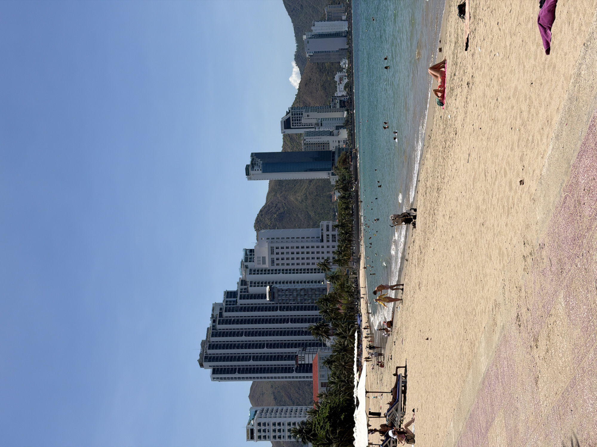 The image depicts a beach in Bac Nha Trang Ward, Vietnam. Tall buildings line the coast, rising against the backdrop of a mountain. People are scattered along the sandy beach and in the water. Some people are swimming in the sea. Others are sunbathing or walking on the beach. Beach chairs and umbrellas are placed near the edge of the sand.