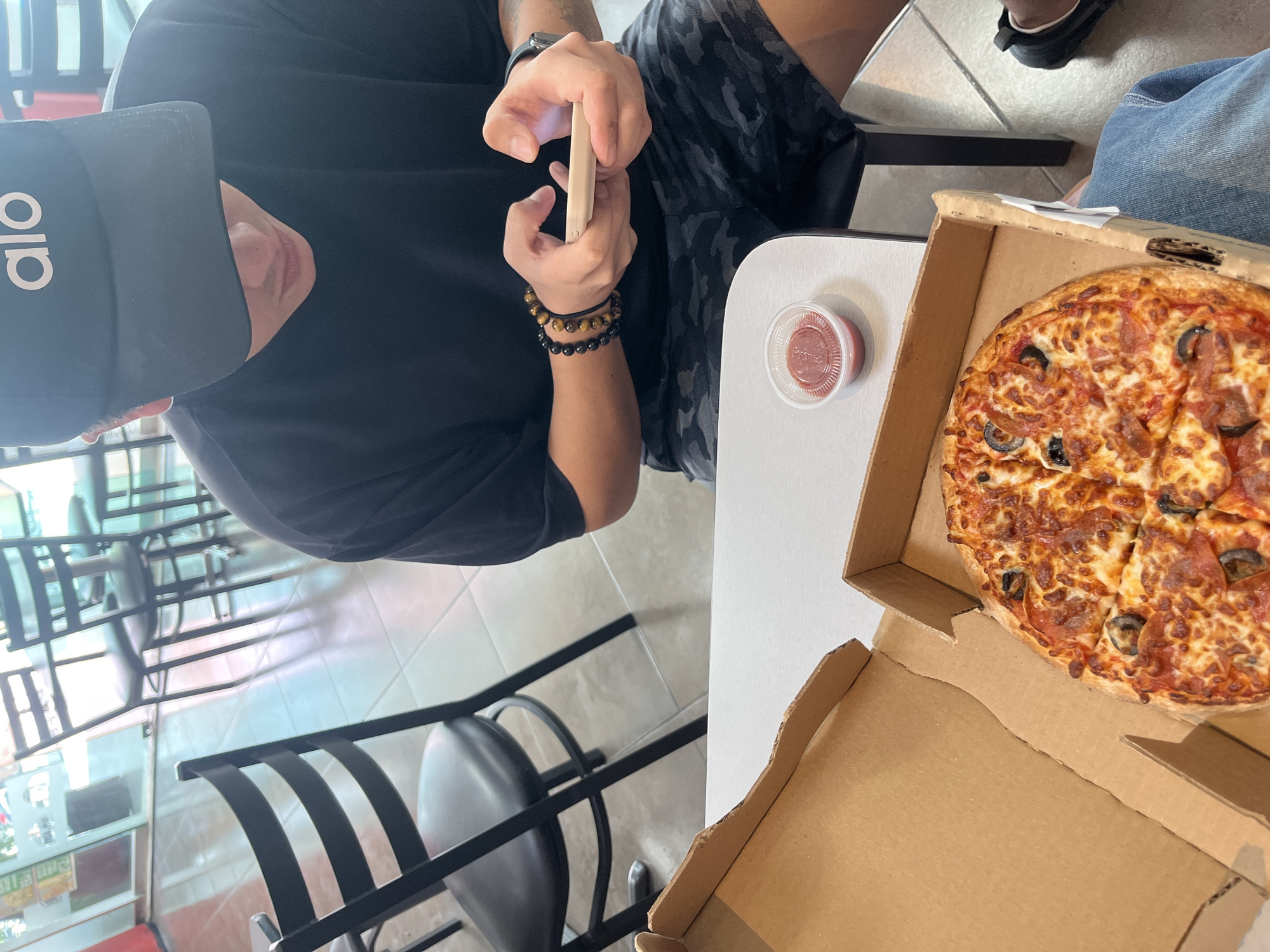 The image depicts an indoor scene at a casual dining establishment in San Bernardino Tlaxcalancingo, Mexico. In the lower left foreground, a person's lower body is visible, wearing blue jeans and black athletic shoes, with an open brown cardboard pizza box resting on their lap. Inside the box is a medium-sized, round pizza topped with pepperoni, black olives, and melted cheese. To the right, a second individual is seated at a light-colored table, holding a light-colored smartphone horizontally with both hands, actively interacting with the screen. This person is wearing a black t-shirt, camouflage shorts, and a black baseball cap with "alo" in white text, which largely obscures their face. Multiple beaded bracelets, including black and tiger's eye beads, are visible on their left wrist, along with a dark smartwatch on their right wrist. A small, clear plastic cup containing red sauce is positioned on the table between the two individuals. The floor consists of light-colored square tiles, and the background features large glass windows or doors offering a blurred view of an outdoor area with natural light and hints of outdoor seating.