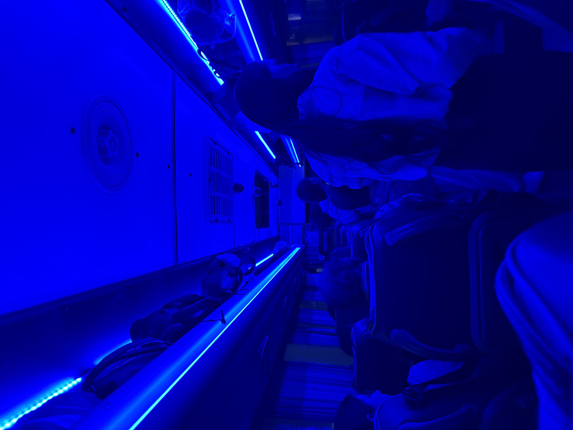 The image depicts the interior of a bus, illuminated by vibrant blue LED lighting strips that run along the ceiling. Several passengers are visible, seated and facing forward, their features somewhat obscured by the low light and blue cast. In the foreground, a person's back and shoulder are visible, wearing a light-colored jacket. Further down the aisle, other passengers are seen from behind, some with dark, curly hair. Luggage is stowed on overhead racks. The bus appears to be in motion or parked, as suggested by the arrangement of seats and the interior lighting. The scene is devoid of any specific landmarks or indications that would place it in Kazan, Russia.