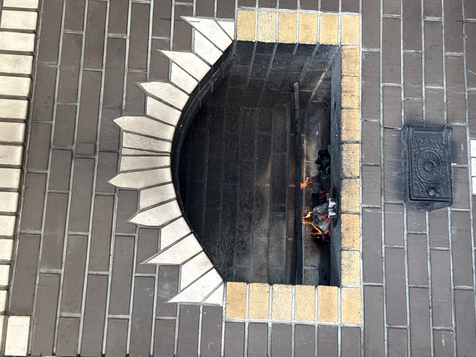 The image depicts a brick outdoor fireplace, seen from above. The fireplace has a rectangular firebox surrounded by lighter-colored bricks. Inside the firebox, wood is burning, with visible flames. The firebox is enclosed on three sides by brickwork. The front of the fireplace has a curved opening with a decorative white brick arch. The surrounding ground is paved with grey bricks. A small, ornate metal door is set into the brickwork to the right of the firebox. The specific location in Солонцы, Russia, cannot be determined from the image.