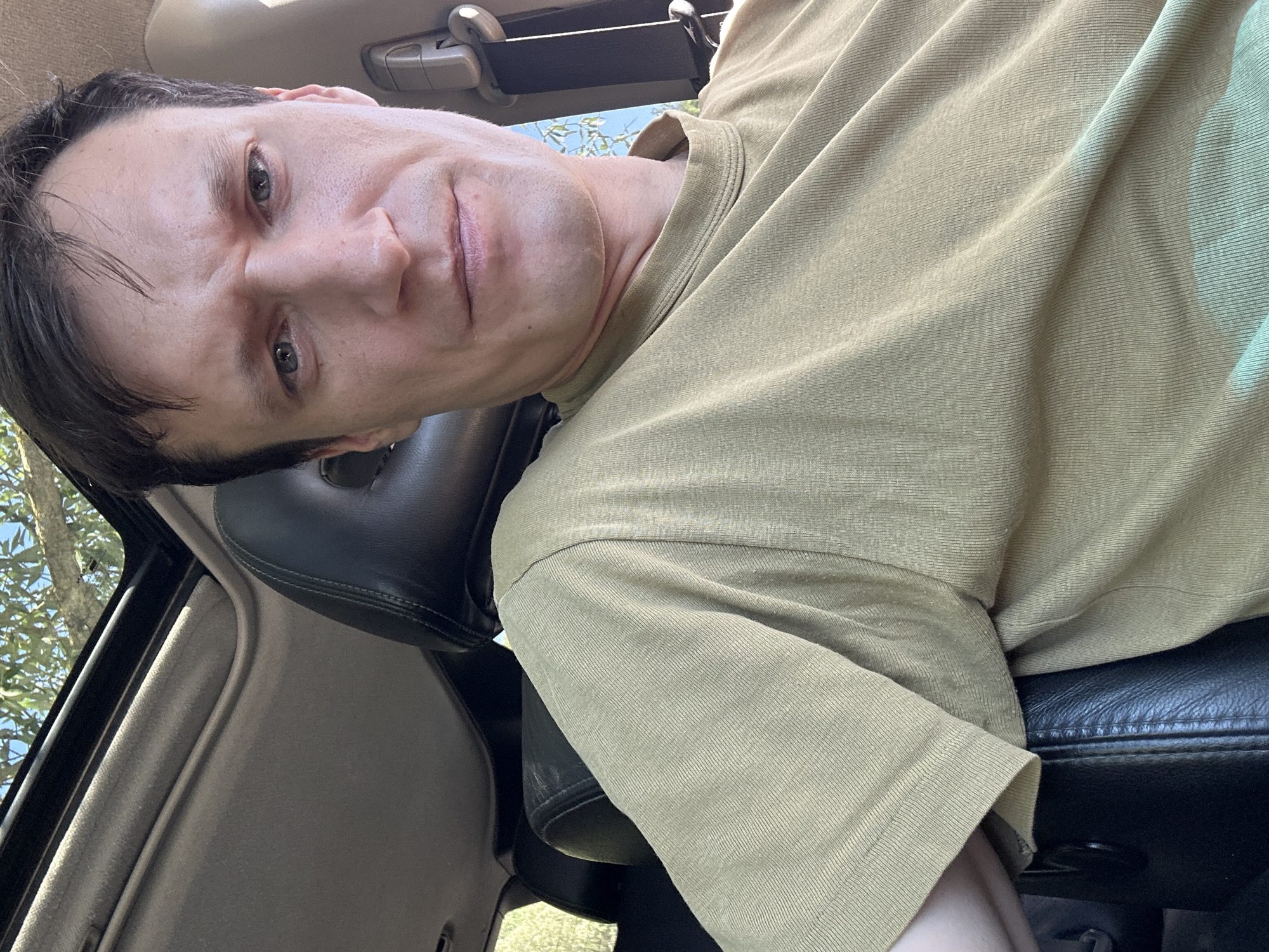 A medium close-up selfie shows a Caucasian man with short, dark hair and blue eyes. He is sitting in the passenger seat of a car, as indicated by the interior upholstery and sunroof visible in the background. The man is wearing a tan, short-sleeved shirt. His facial expression is neutral, and he is looking directly at the camera. The scene is outdoors, as evidenced by the sunlight streaming through the sunroof and the foliage visible above. There is no specific indication of the location within the city of Makiv, Ukraine.