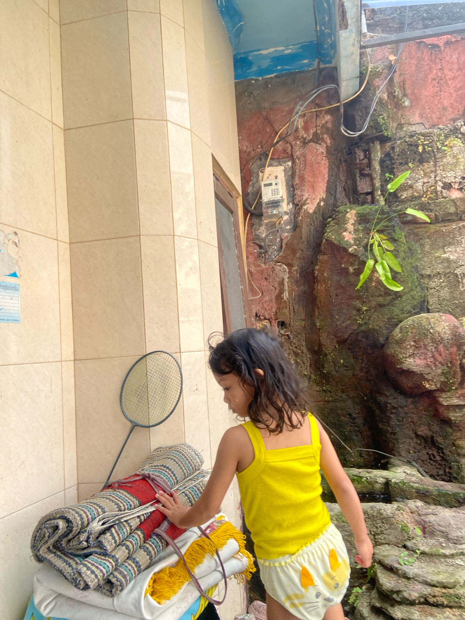 In Gelam, Indonesia, a young girl with dark, wavy hair is captured in a candid moment, standing near a tiled wall. She wears a bright yellow tank top and light-colored shorts adorned with a cat pattern. Her right hand is extended, touching a stack of rolled-up mats and blankets, one of which is predominantly red and blue striped. A badminton racket rests against the mats. The background reveals a rough, moss-covered stone wall with exposed wiring and an electrical meter box mounted on it. The scene suggests an outdoor setting, possibly a backyard or alleyway, with natural elements like plants and moss contributing to the rustic ambiance.