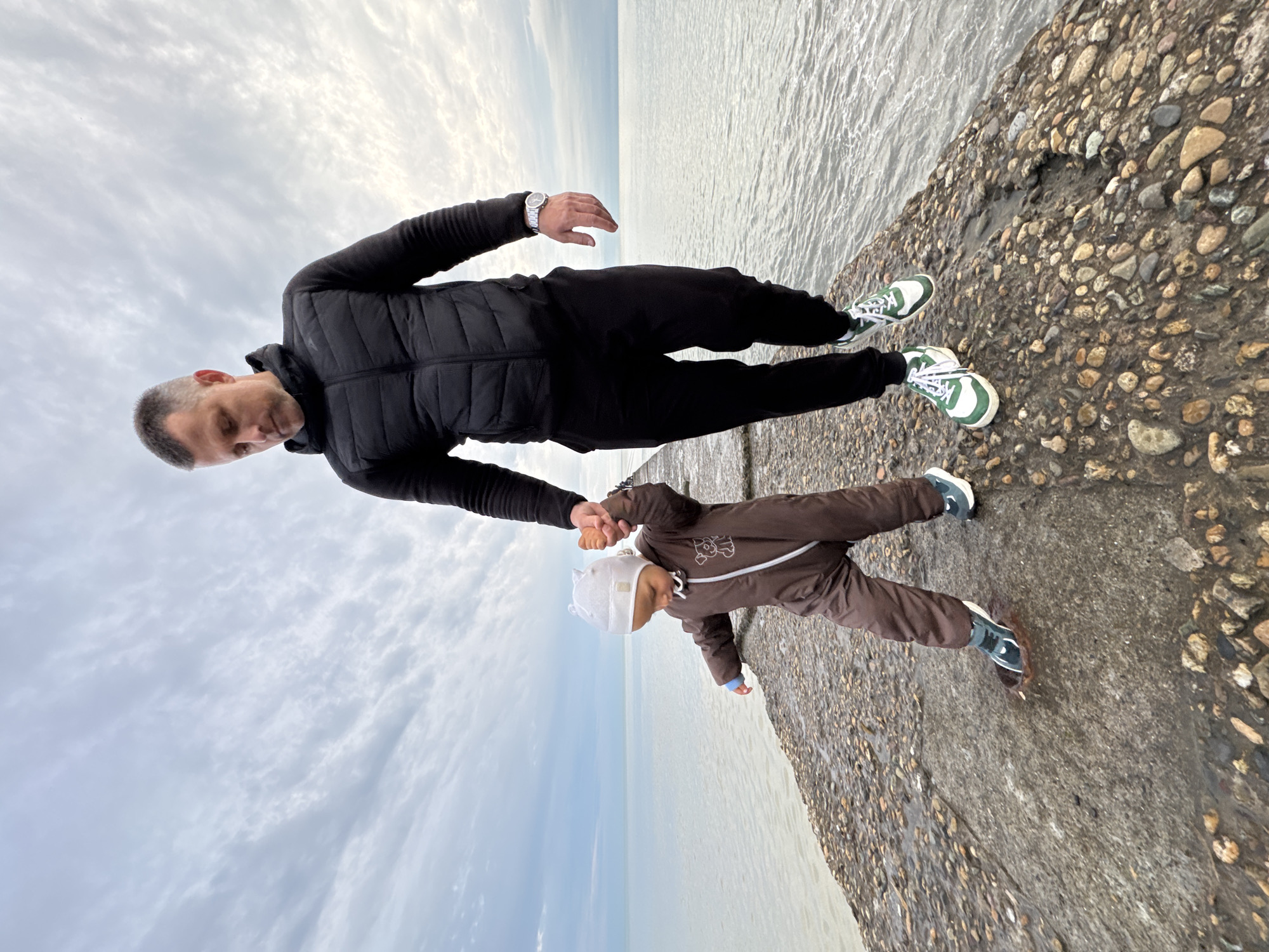 A full-length outdoor photograph, initially rotated 90 degrees clockwise, captures a man and a young child walking or standing on a concrete pier by the sea under an overcast sky. Once correctly oriented, the man is seen on the left, wearing a black puffer jacket, black pants, and green and white sneakers. A silver watch is visible on his left wrist. He has short, graying hair and is looking down at the child, holding their hand.
The child, positioned to the man's right, is dressed in a brown one-piece suit with a white zipper and a light-colored bear motif on the chest, a white hat with small ear-like features, and dark sneakers. They are standing on a textured concrete surface embedded with pebbles. To the right of the subjects, calm water reflects the cloudy sky, extending to a faint horizon line. The sky is largely covered by soft, light gray and white clouds. The attire suggests cool weather on an overcast day. The scene depicts a quiet, protective interaction between the adult and child at a coastal location, identified as Sochi, Russia, likely by the Black Sea. No visible text is present.