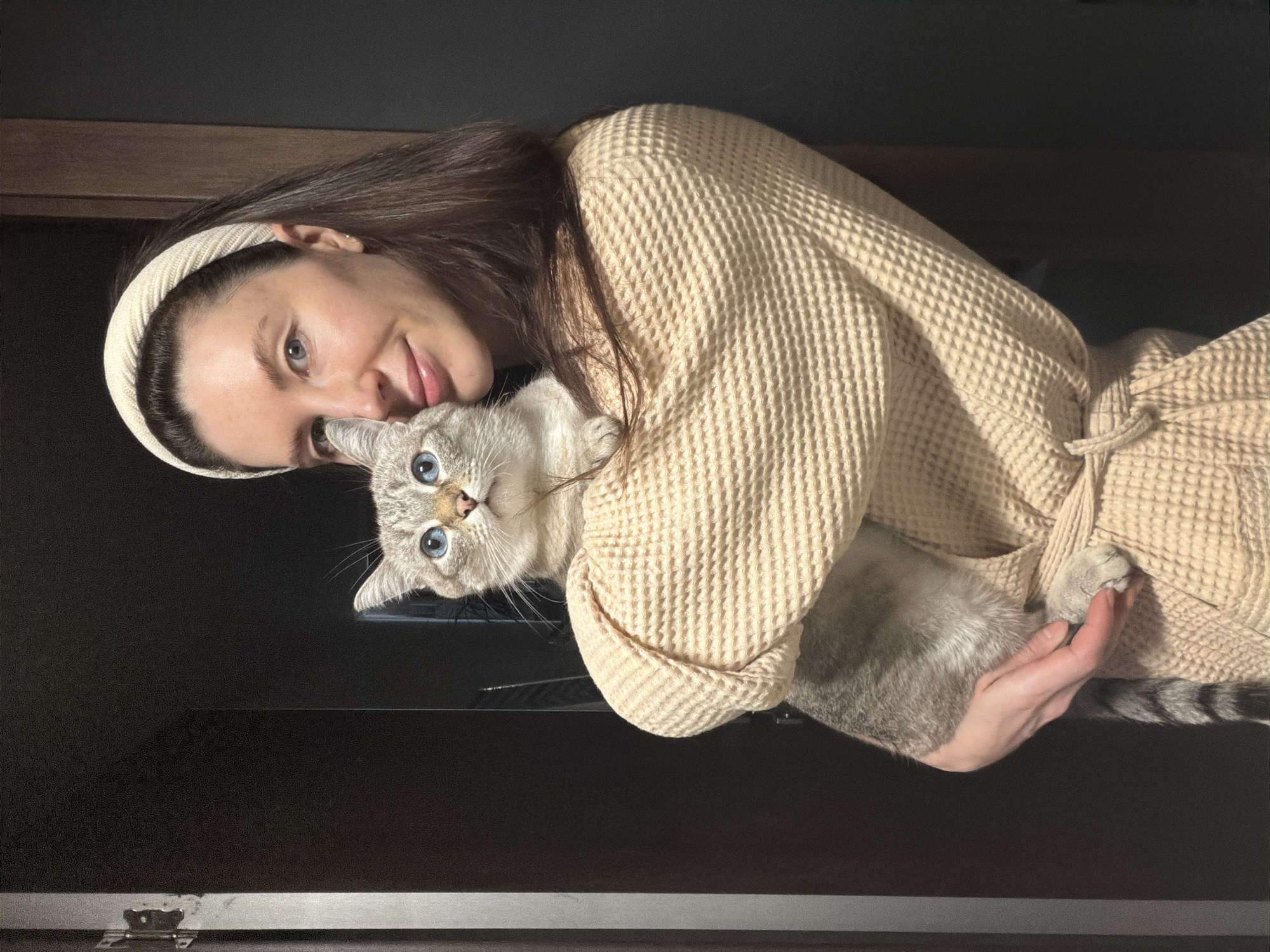 A woman with long brown hair, wearing a cream-colored waffle-textured robe and a matching headband, is holding a light gray tabby cat with striking blue eyes. The woman is smiling and looking at the camera, while the cat is also looking directly at the camera. The background is a dark, possibly black wall. The photo appears to have been taken indoors, potentially in Perm, Russia.
