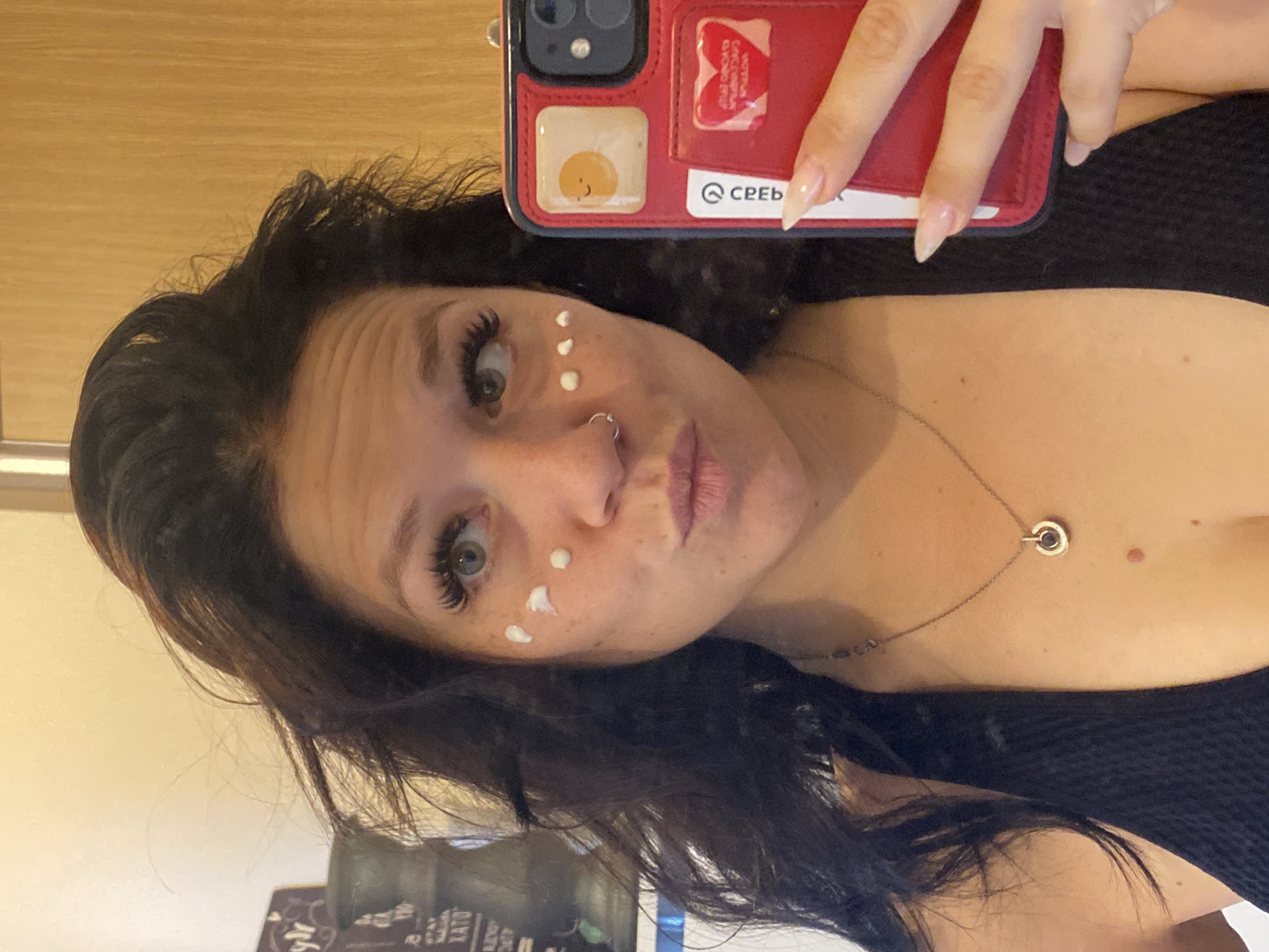 A medium shot shows a woman holding a red smartphone in her right hand, angled to capture her reflection in the screen. The woman has dark, wavy hair that frames her face and is wearing a black top. She is applying white cream to her face, with dots of cream visible on her cheeks. A silver necklace with a circular pendant is around her neck. The background shows a blurred interior with wooden paneling and decorative items on a shelf. The image is taken indoors, likely in a residential setting.