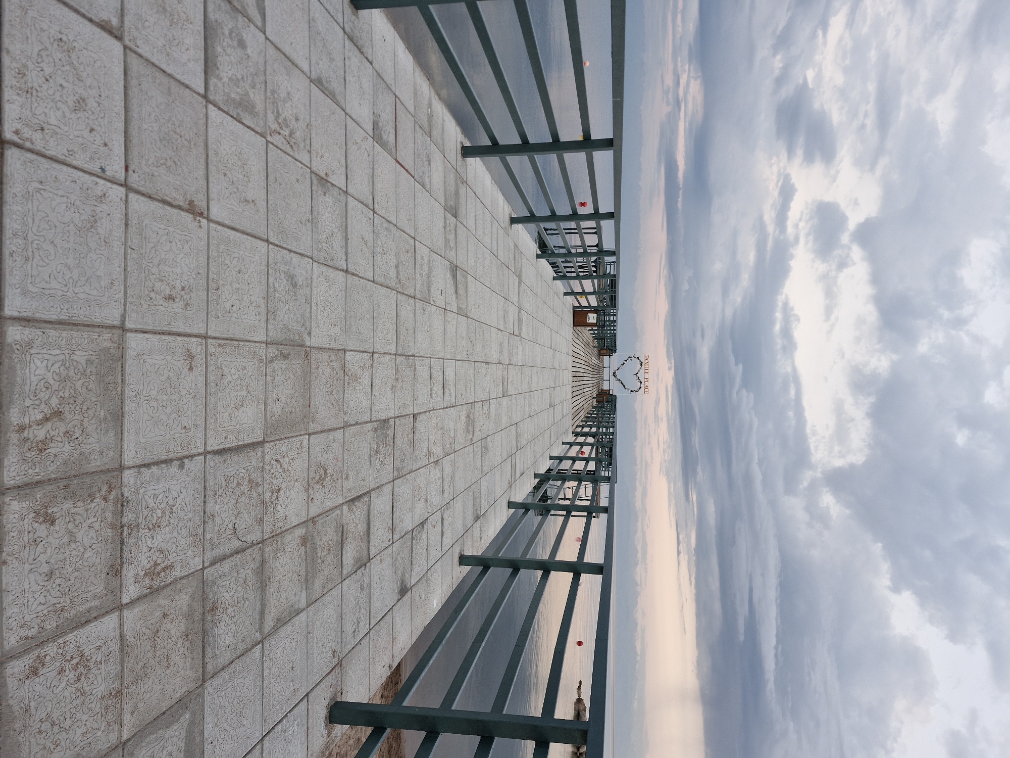 The image depicts a long, paved pier extending out over a body of water. The pier is flanked by a dark green metal railing. The paving stones are square and light-colored, with a decorative pattern on many of them. In the distance, at the end of the pier, a large heart-shaped structure with lights is visible, along with the words "FAMILY PLACE". The sky is overcast with dramatic, cloudy formations, and the water is calm. The overall scene suggests a public waterfront area, possibly a tourist attraction or recreational spot. The location is identified as Bulan-Segettu, Kyrgyzstan. There are no people or animals visible in the image. The environment is serene and appears to be either early morning or late evening due to the soft lighting.