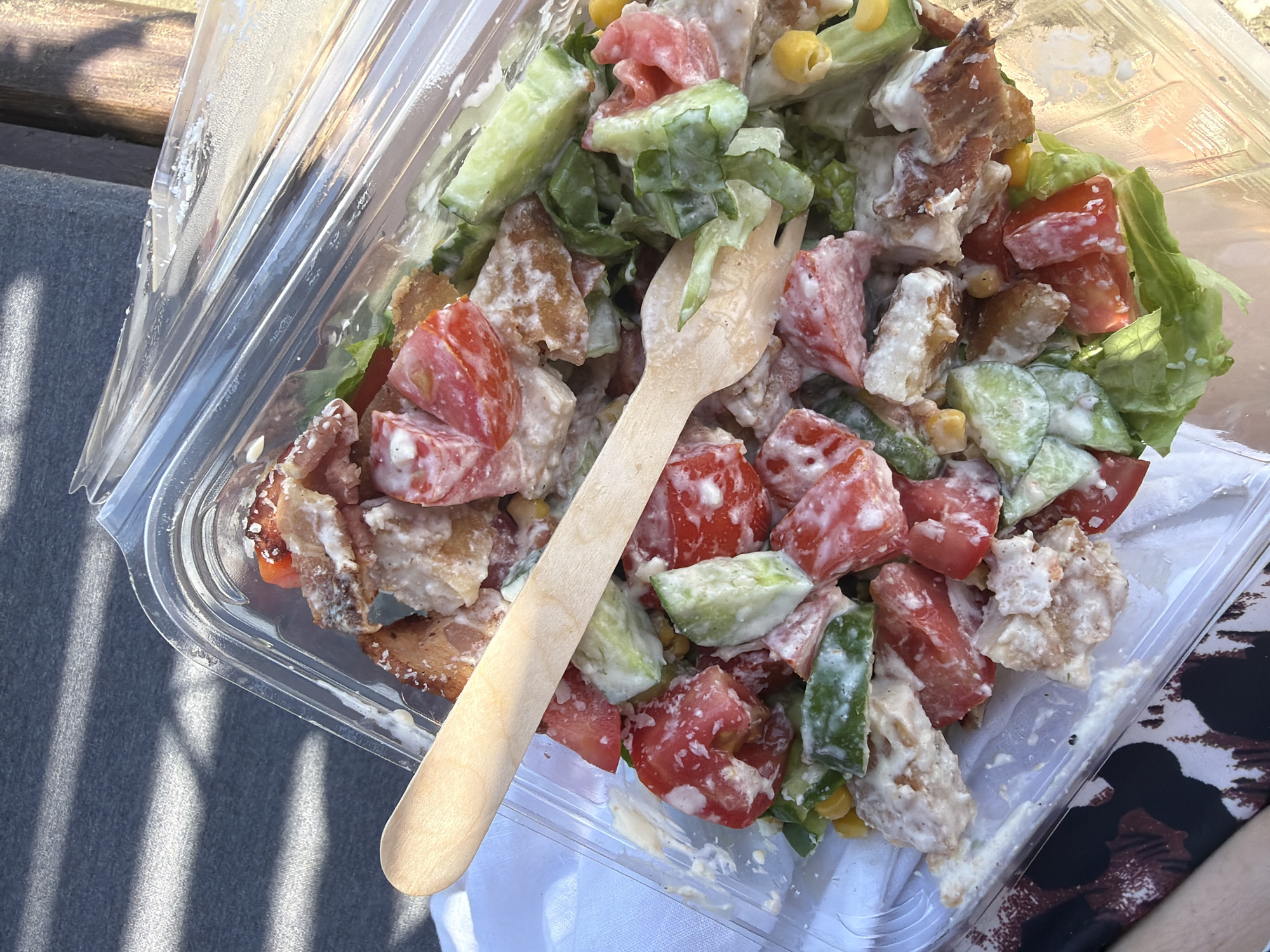 An overhead, close-up shot captures a salad served in a clear plastic container, resting on a textured grey surface. The salad is a vibrant mix of chopped tomatoes, cucumber slices, lettuce, corn kernels, and pieces of fried chicken, all coated in a creamy white dressing. A wooden fork is placed across the top of the salad, with its tines partially submerged in the ingredients. The background shows dappled sunlight casting shadows, suggesting an outdoor setting. The scene is located in Meneou, Cyprus.