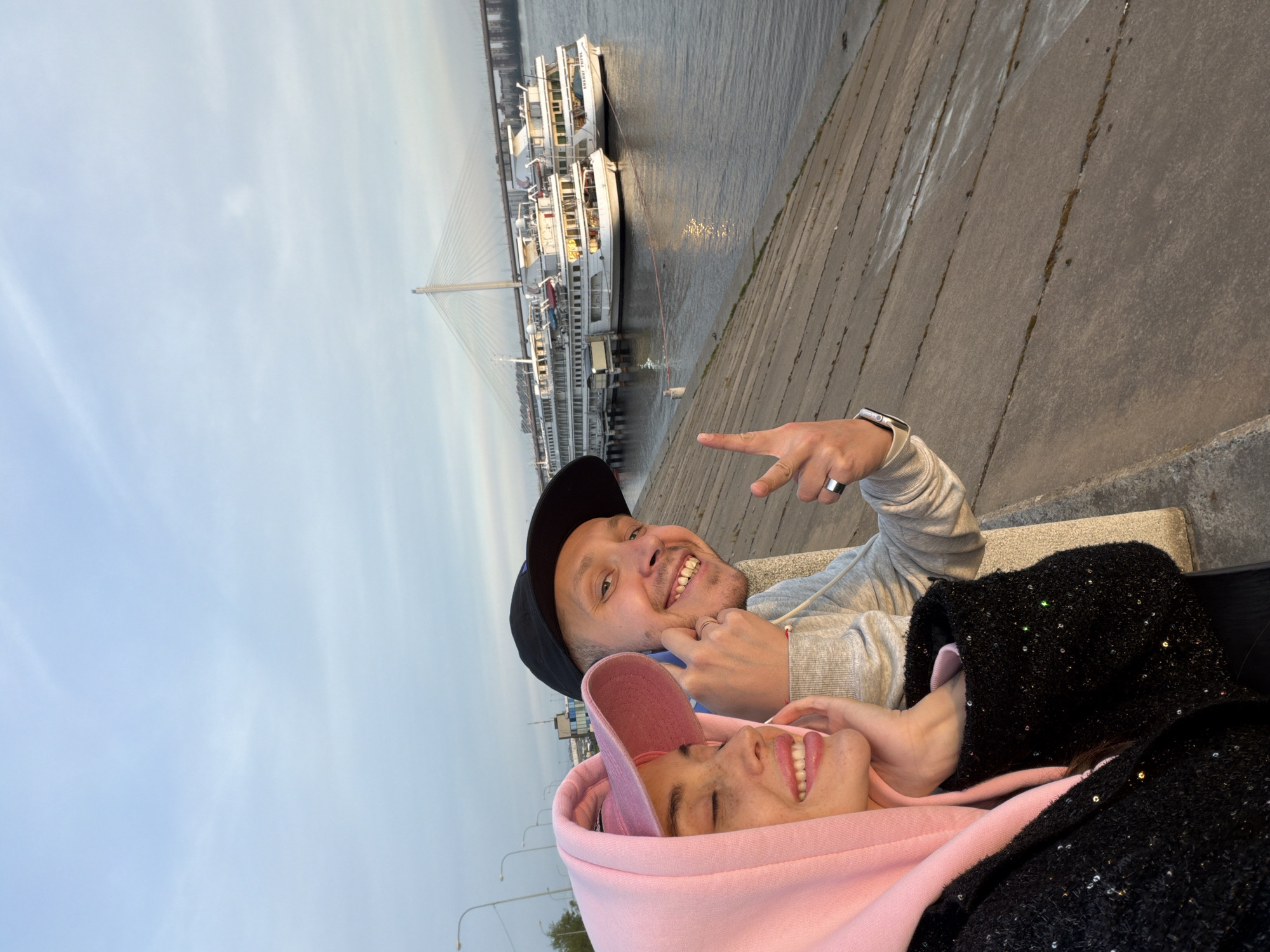 Two people are reclining on a concrete bench near a body of water. The person on the left is wearing a pink hooded sweatshirt. The person on the right is wearing a gray sweatshirt and a black baseball cap and pointing at something in the distance. Both are smiling. A bridge and several boats are visible in the background. The scene is in Saint Petersburg, Russia.