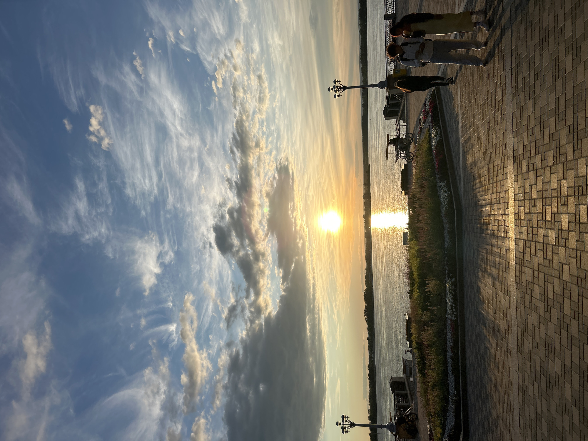 The image depicts a picturesque sunset scene on a paved promenade alongside a body of water. Several people are visible walking along the promenade, silhouetted against the bright light of the setting sun. Some are gathered together, possibly in conversation, while others walk individually. Objects present include lampposts, a bicycle, and flowering plants in a garden bed along the edge of the water. The sky is a dramatic mix of blue and white clouds, with streaks of orange and yellow as the sun dips towards the horizon. The water reflects the golden hues of the sunset, creating a shimmering path across its surface. The paving stones on the promenade cast long, diagonal shadows, indicating the low angle of the sun. The overall atmosphere is serene and peaceful, characteristic of a pleasant evening. The location is identified as Novokuybyshevsk, Russia.