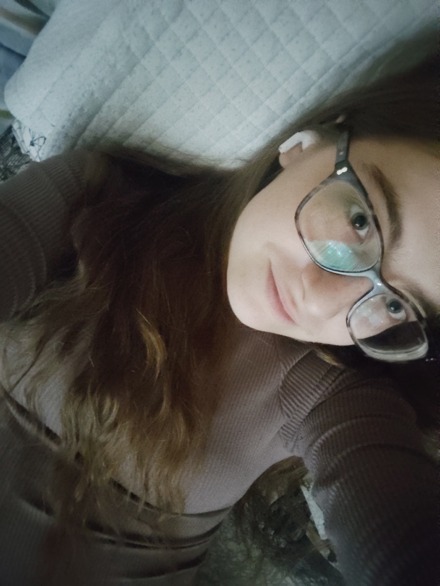 A woman with brown hair is lying down, possibly on a bed or sofa, with a quilted cushion behind her head. She is wearing glasses and a close-fitting, ribbed top. She has an earbud in her right ear. The image appears to be a selfie taken from a high angle. No information indicates the location as Sofia, Bulgaria.