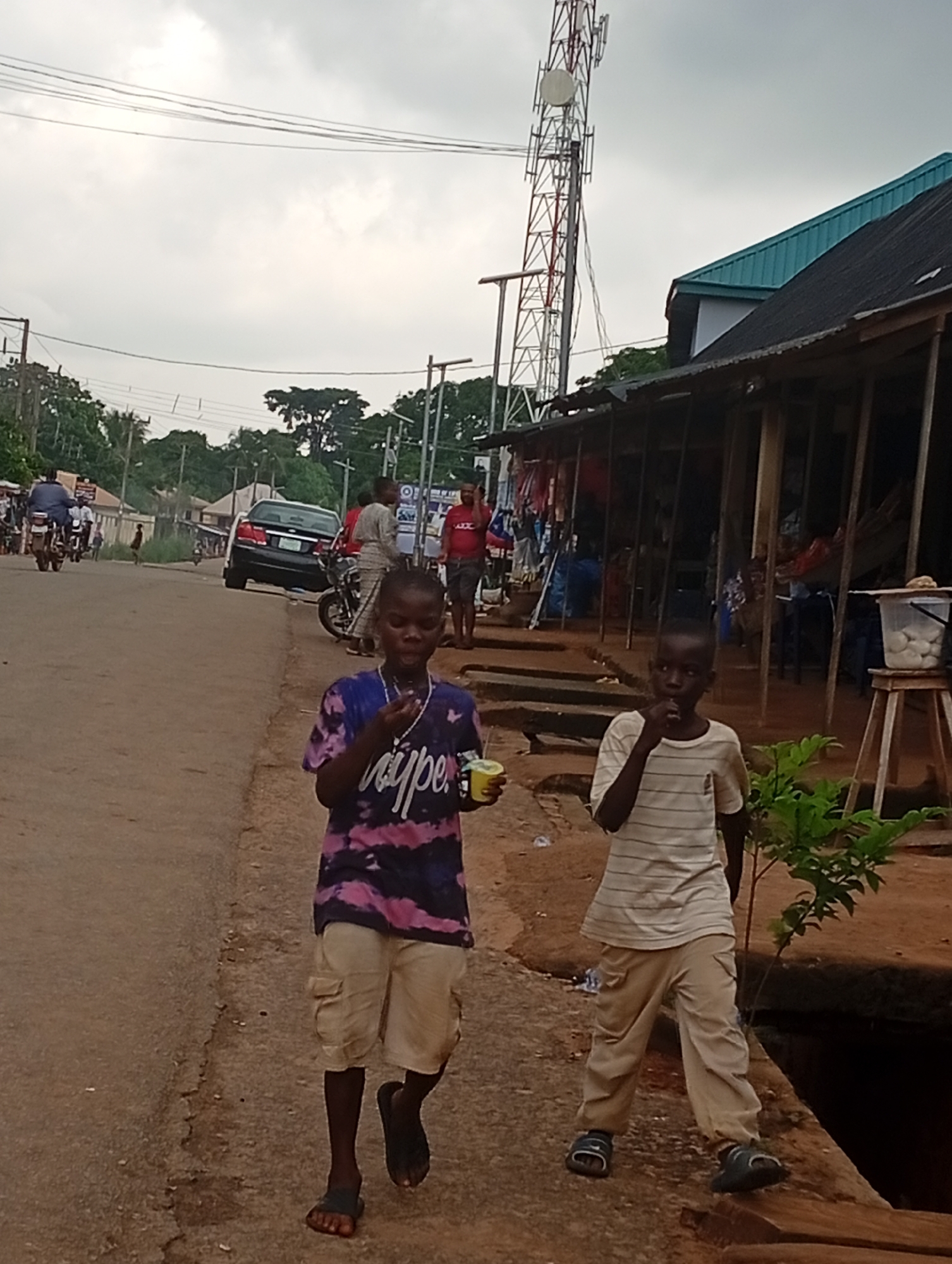 This is a full-shot, eye-level, outdoor photograph taken in Nsugbe, Nigeria. The scene depicts a dirt road lined with businesses and a telecommunications tower in the background. Two young boys are walking towards the camera on the right side of the road. The boy in the foreground on the left is wearing a purple and pink tie-dye shirt and tan cargo shorts, and he is holding a yellow cup and a white object, possibly food. The boy on the right is wearing a white striped t-shirt and tan pants, and he is bringing his hand to his mouth. Behind them, other individuals and vehicles are visible, including motorcycles and a car. The businesses on the right appear to be open-air stalls with goods displayed. The sky is overcast.