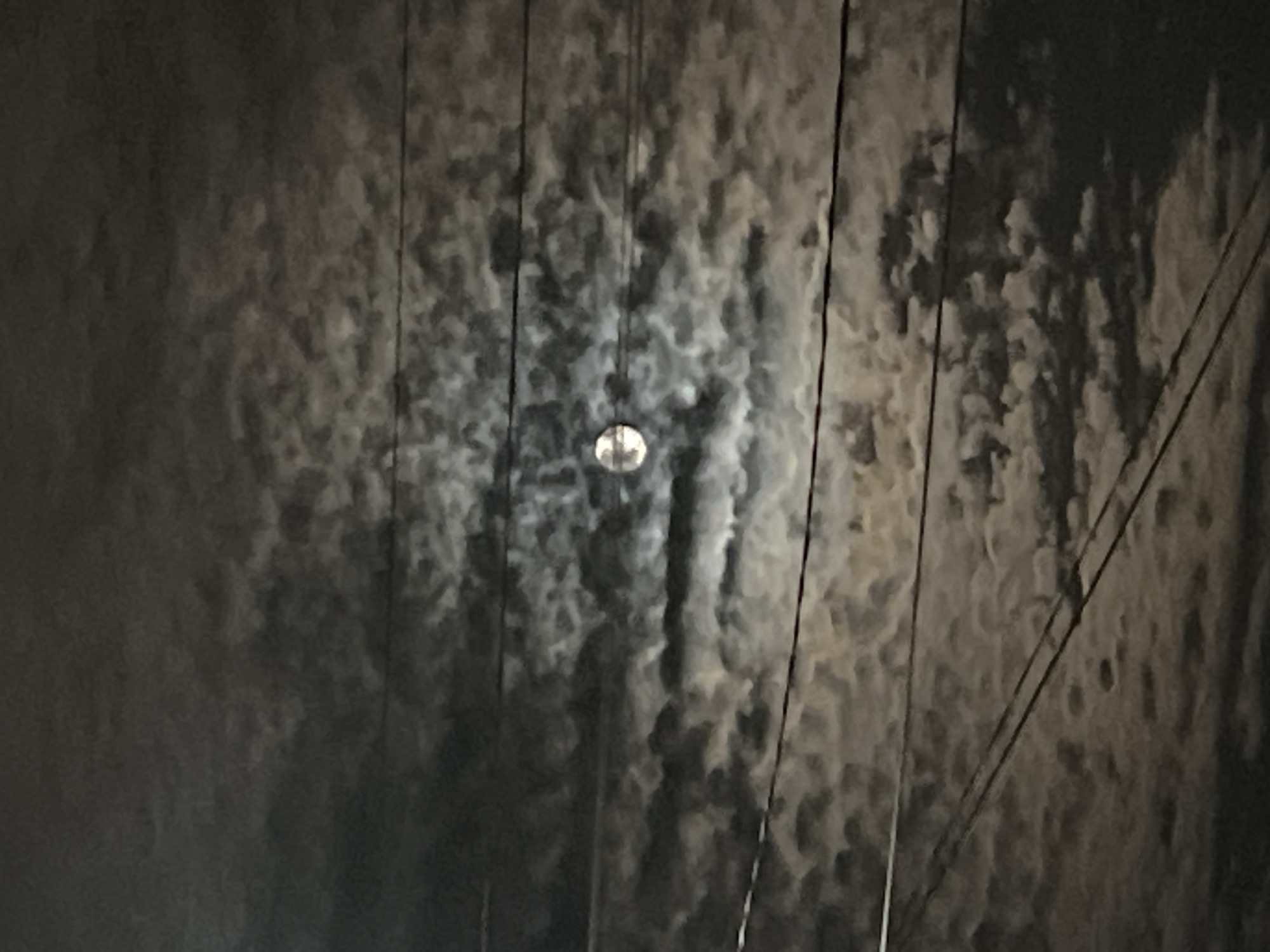 The image depicts a night scene in an urban environment. The focal point is a bright, circular object, possibly a street light or the moon, visible through a textured surface that appears to be a wall or fence made of dark, mottled material, potentially wood or metal with weathering. Multiple dark, thin lines, likely electrical or telephone wires, run diagonally across the scene, adding depth and indicating urban infrastructure. The overall ambiance is dark and somewhat indistinct, suggesting low light conditions. The location of the scene is Unknown, Unknown.