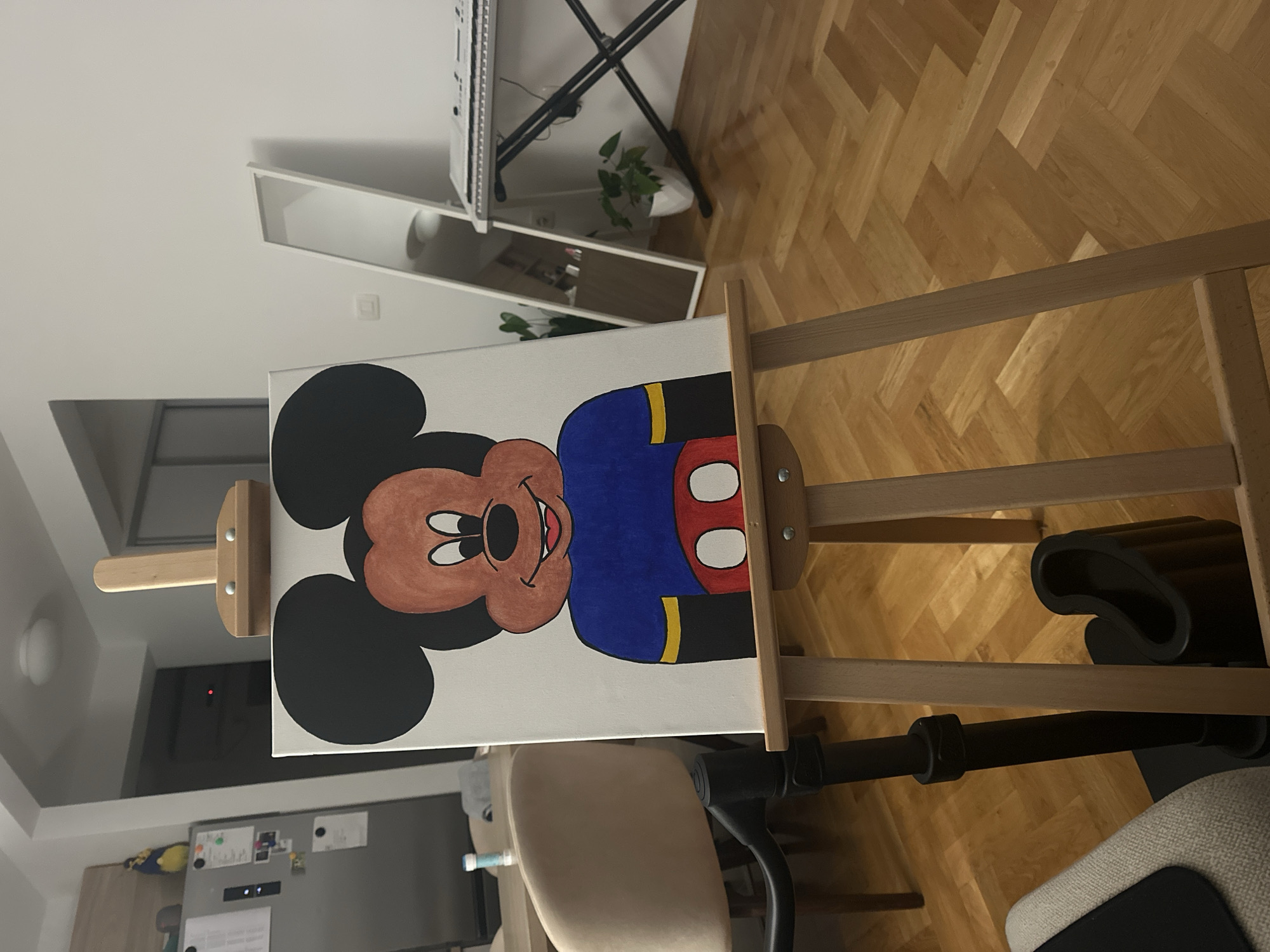 The image shows a painting of Mickey Mouse on a canvas, propped up on a wooden easel. The painting features Mickey Mouse in a blue outfit with a red and white belt, outlined in yellow. The background of the painting is white.
The easel is located indoors. The environment includes a hardwood floor, a mirror, a keyboard stand, a plant, a chair and part of a refrigerator.
The image does not show any indication of being in Budva, Montenegro. There are no people visible.