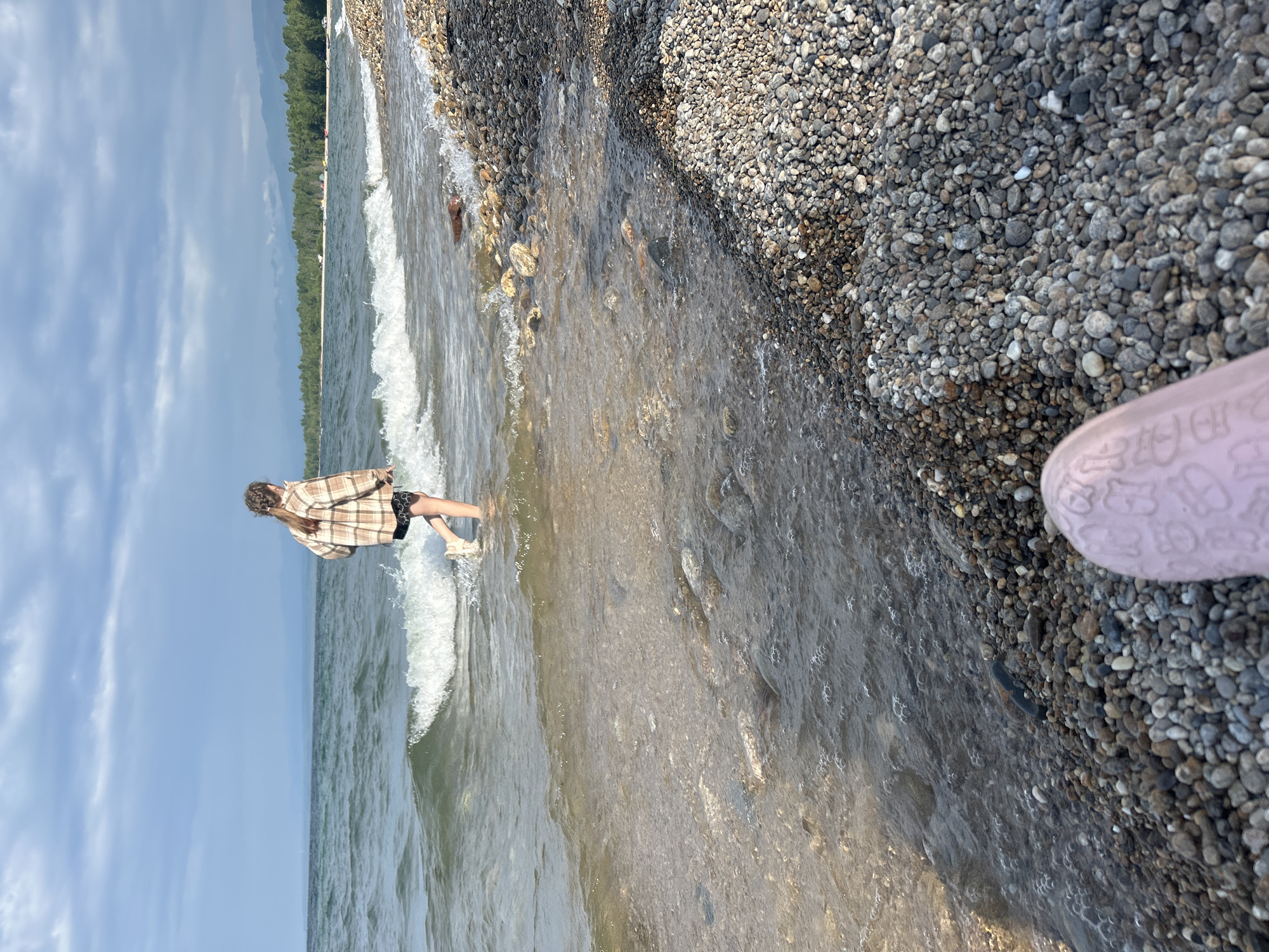 A person is walking in shallow water near a pebble beach. The person is wearing a plaid shirt and shorts. The sky is cloudy, and there are mountains in the distance. The water is calm with small waves breaking on the shore. The beach is covered in smooth, multi-colored pebbles. The foreground shows a pink object that appears to be a shoe or slipper. The scene is likely located at Lake Baikal near Tolbazikha, Russia.