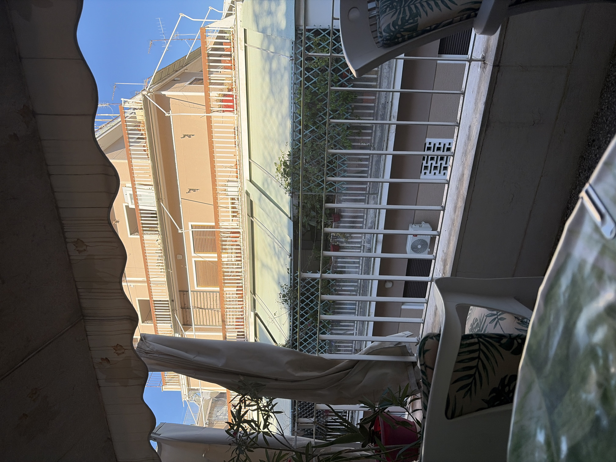This image captures a view from a balcony in Athens, Greece, looking out onto an apartment building and the sky. The balcony has a white railing and is partially covered by a beige awning. In the foreground, the edge of a table with a green patterned tablecloth is visible, along with a white chair with cushions featuring a palm leaf pattern. To the left, plants in a red pot are present. Beyond the balcony, a multi-story apartment building with peach-colored walls and white railings on its balconies is depicted. Several windows with brown shutters are visible. A green awning extends across part of the building's facade. In the distance, the bright blue sky is clear. The scene appears to be set in a residential area, likely during daylight hours.