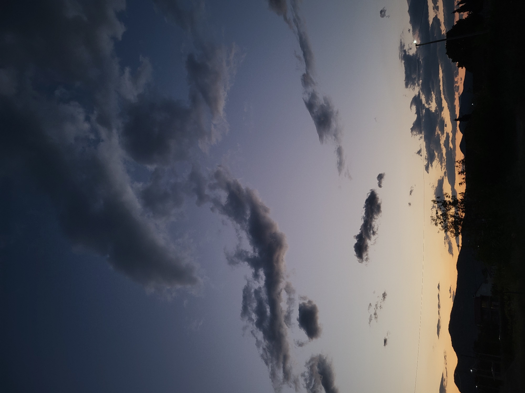 This image captures a twilight landscape scene in Mahala, Montenegro, characterized by a dramatic sky and a silhouetted horizon. The sky exhibits a striking gradient, transitioning from a deep indigo or dark blue in the upper left to a lighter, more ethereal blue-violet across the middle, and finally to a warm, glowing orange and yellow near the horizon on the lower right, indicative of either sunset or twilight.

Numerous dark, textured clouds are scattered throughout the sky, ranging from large, dense formations in the darker blue areas to more elongated, wispy streaks against the lighter sky. These clouds add depth and a sense of movement to the expansive sky.

The horizon line, visible on the lower right portion of the image, features a silhouette of undulating mountains or hills. In the immediate foreground, also in silhouette, are outlines of trees and what appears to be a utility pole with a faint light illuminating its tip. A small, indistinct structure, possibly a building or house, is also faintly visible in the distance, nestled against the mountains.

The overall mood is serene and atmospheric, showcasing the natural beauty of the landscape as daylight fades. No people or visible text are present in the image.